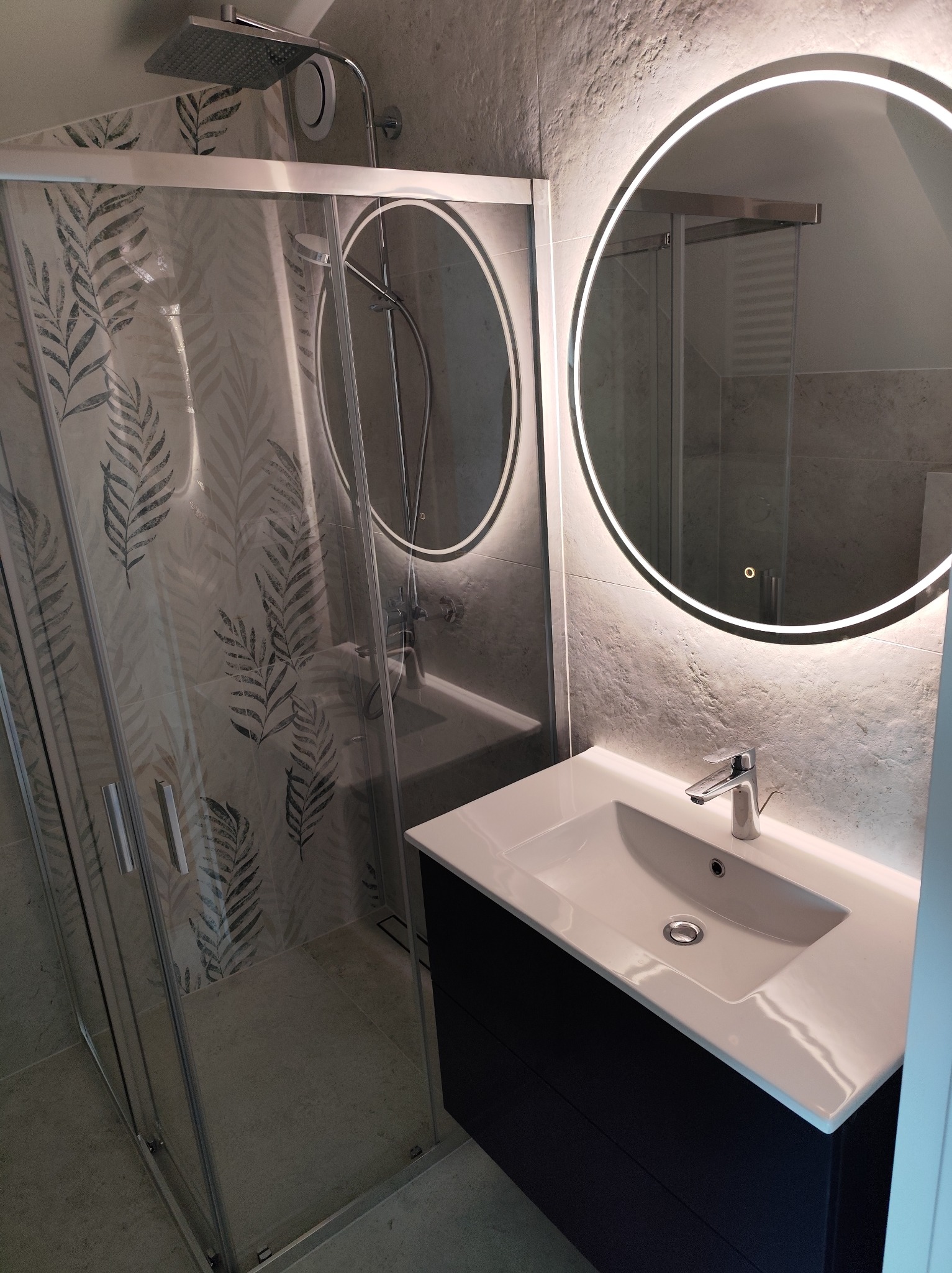 Nowoczesna łazienka z kabiną prysznicową w liście i okrągłym lustrem z podświetleniem LED nad umywalką. Szare płytki imitujące kamień.