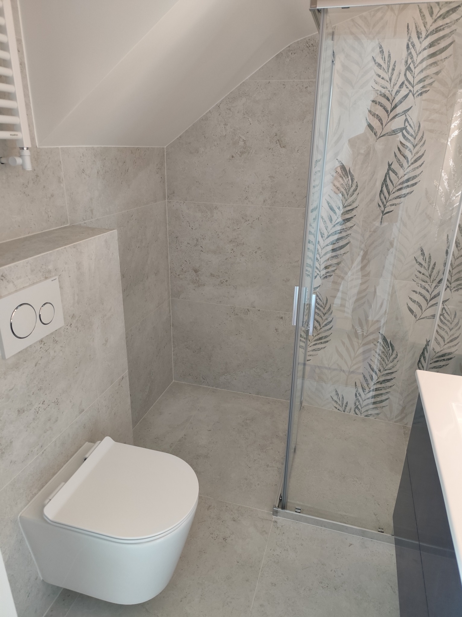 Nowoczesna łazienka w szarościach: podwieszana toaleta, kabina prysznicowa z motywem liści, płytki imitujące kamień, grzejnik drabinkowy. Ukośny sufit.