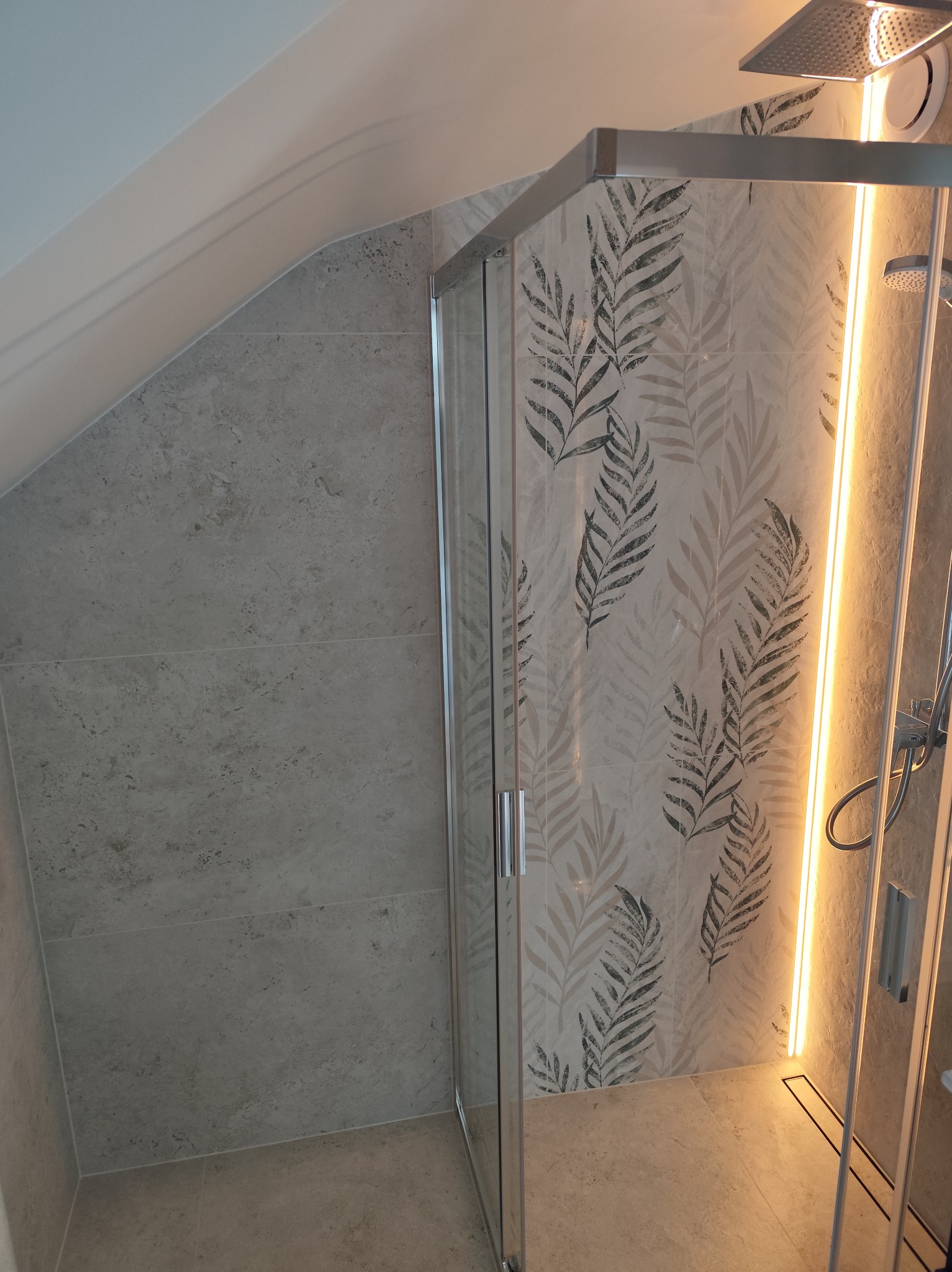 Nowoczesna kabina prysznicowa z szarymi płytkami imitującymi kamień i dekoracyjnym wzorem liści na ścianie, oświetlona listwą LED. Ukośny sufit.