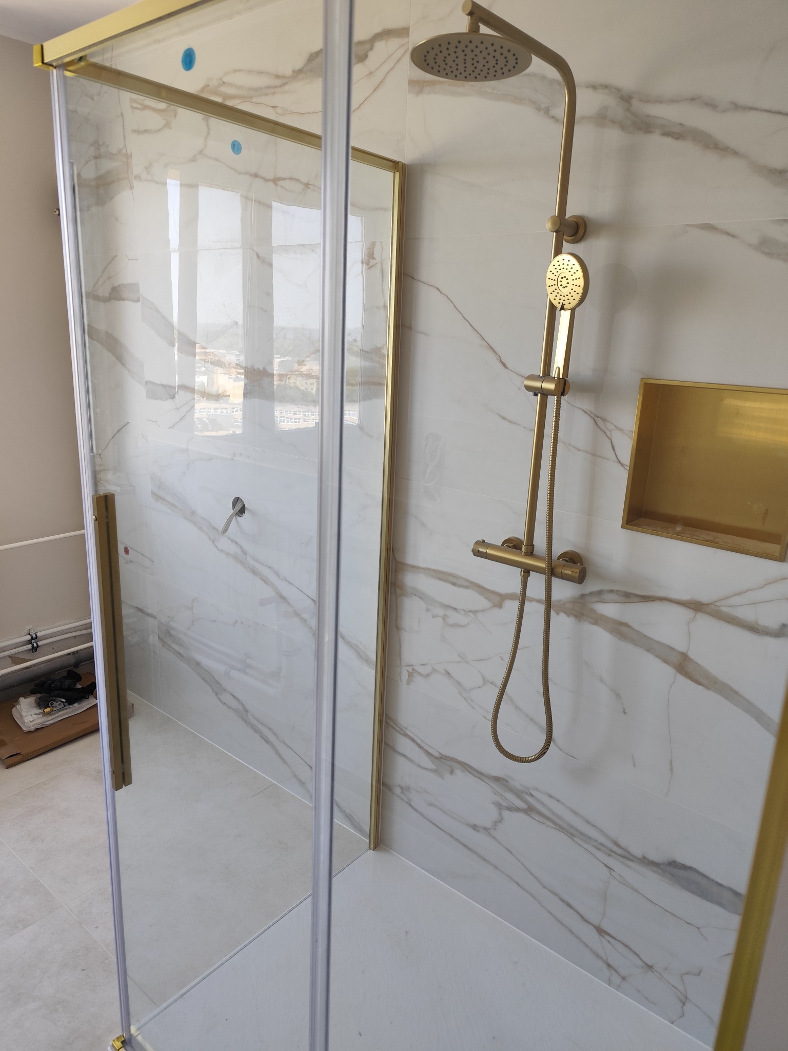 Kabina prysznicowa z złotymi elementami na tle marmurowych płytek. Widoczny zestaw natryskowy i wnęka w ścianie. Nowoczesny design łazienki.