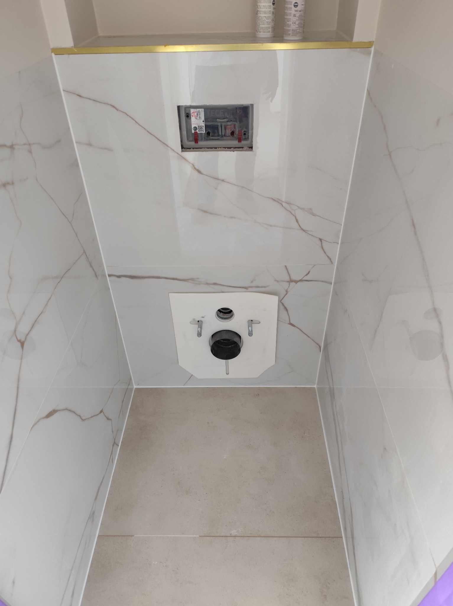 Montaż stelaża podtynkowego WC w łazience w Szczecinie. Białe płytki imitujące marmur z delikatnym, złotym akcentem. Widoczna instalacja przed zabudową.