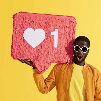 Mężczyzna w okularach trzyma w dłoni różową piniatę w kształcie dymku z sercem i cyfrą jeden na żółtym tle, wyrażający zaskoczenie.