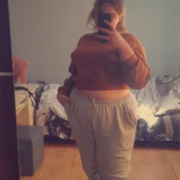 Osoba robiąca selfie w lustrze, ubrana w brązową bluzę i szare spodnie dresowe, widoczne skarpety z motywem liści konopi, na ścianie napis 'Jestem 2005'.