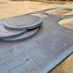 Stos trzech okrągłych, wyciętych metalowych elementów leżących na metalowej płycie z wyciętymi otworami, na drewnianym stole, widoczne ślady obróbki.