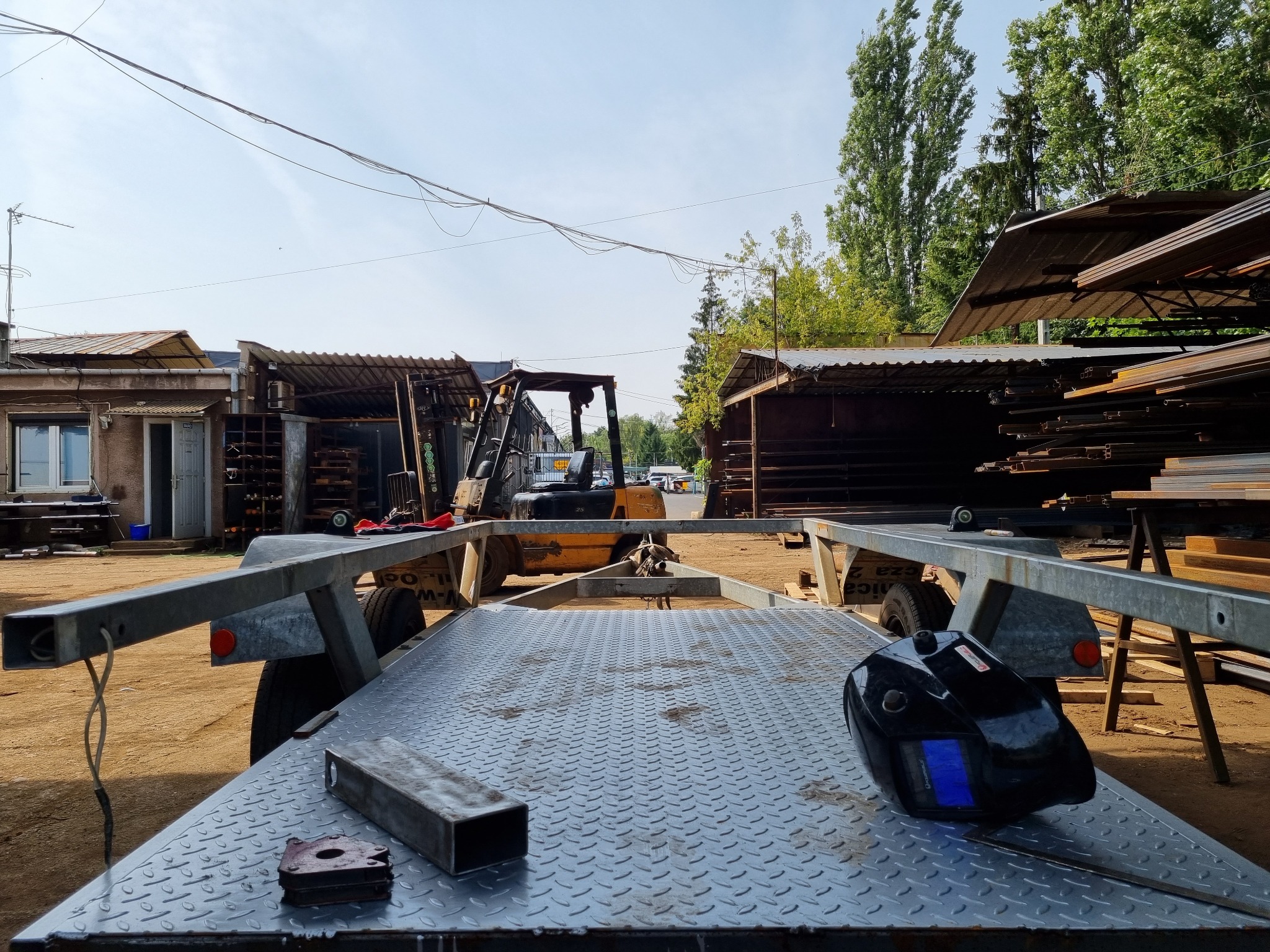 Przygotowany do transportu wózek z metalową konstrukcją, maska spawalnicza i profile stalowe w warsztacie, w tle widoczny wózek widłowy.