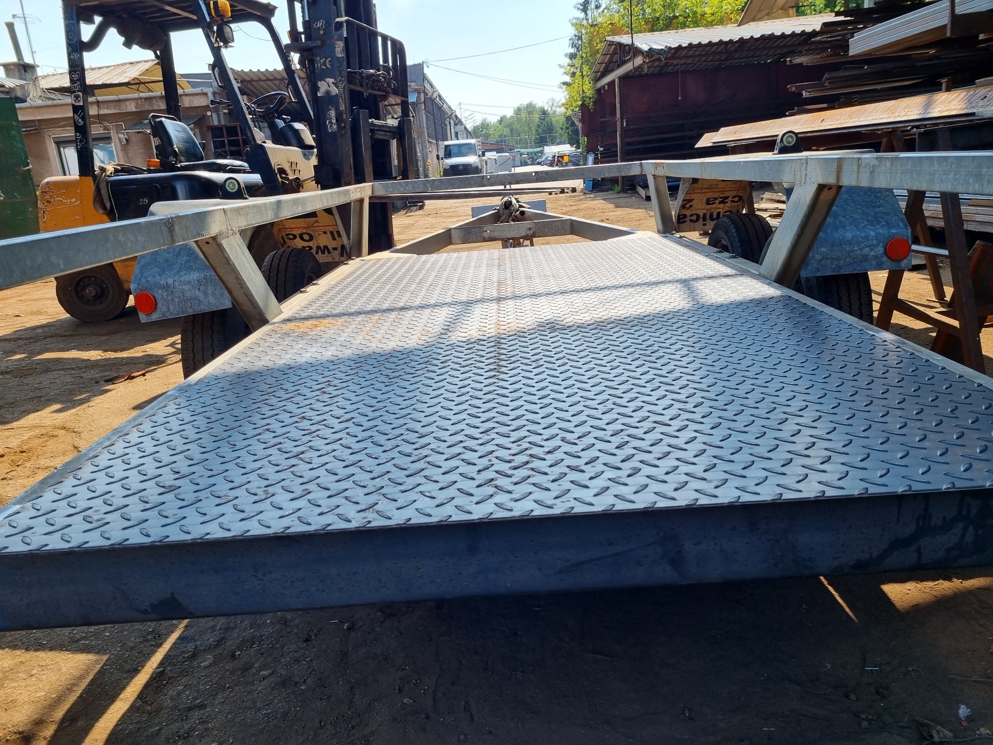 Metalowa przyczepa z blachą ryflowaną na placu budowy, z widocznym wózkiem widłowym i konstrukcjami stalowymi w tle.