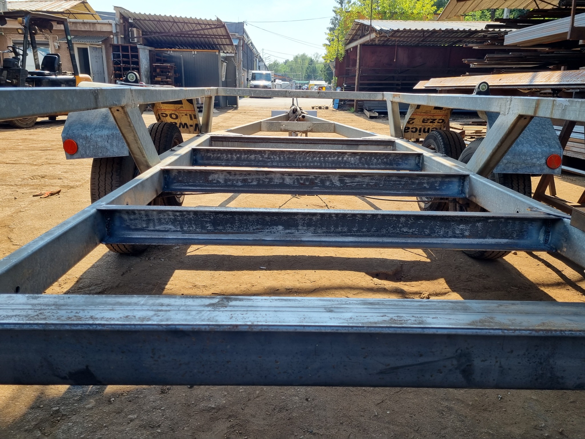 Metalowa rama przyczepy samochodowej z widocznymi spawami i zamontowanymi kołami, ustawiona na placu budowy w słoneczny dzień.