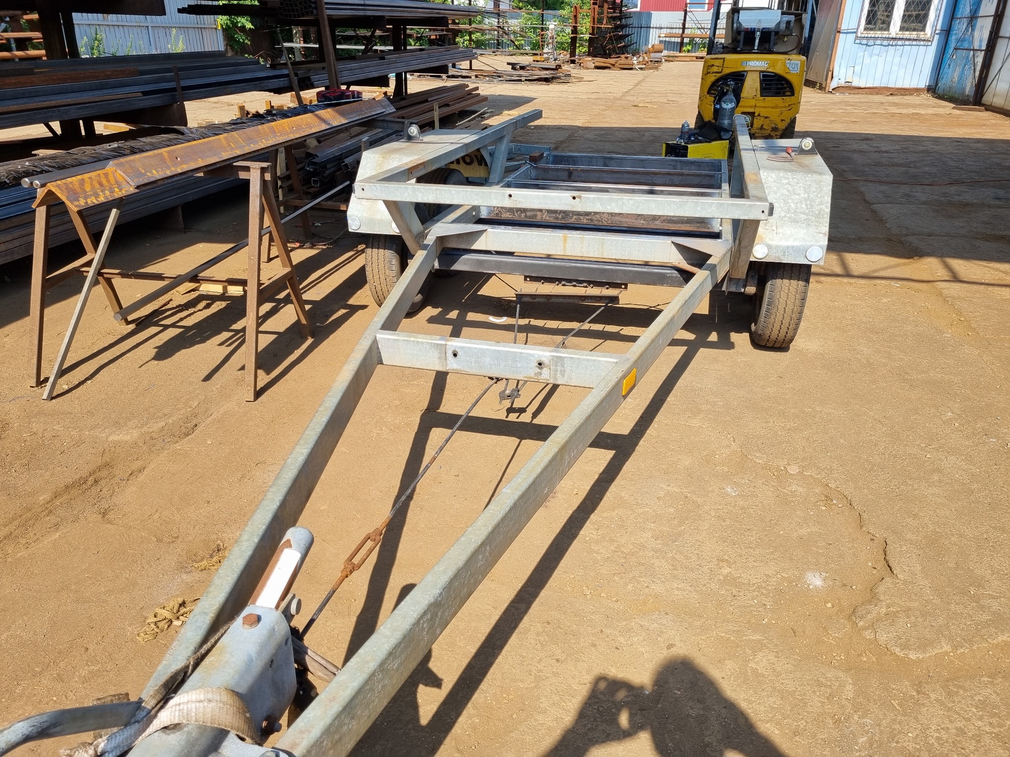 Metalowa konstrukcja przyczepy w warsztacie, widoczne profile stalowe i spawane elementy, w tle składowane materiały i wózek widłowy.