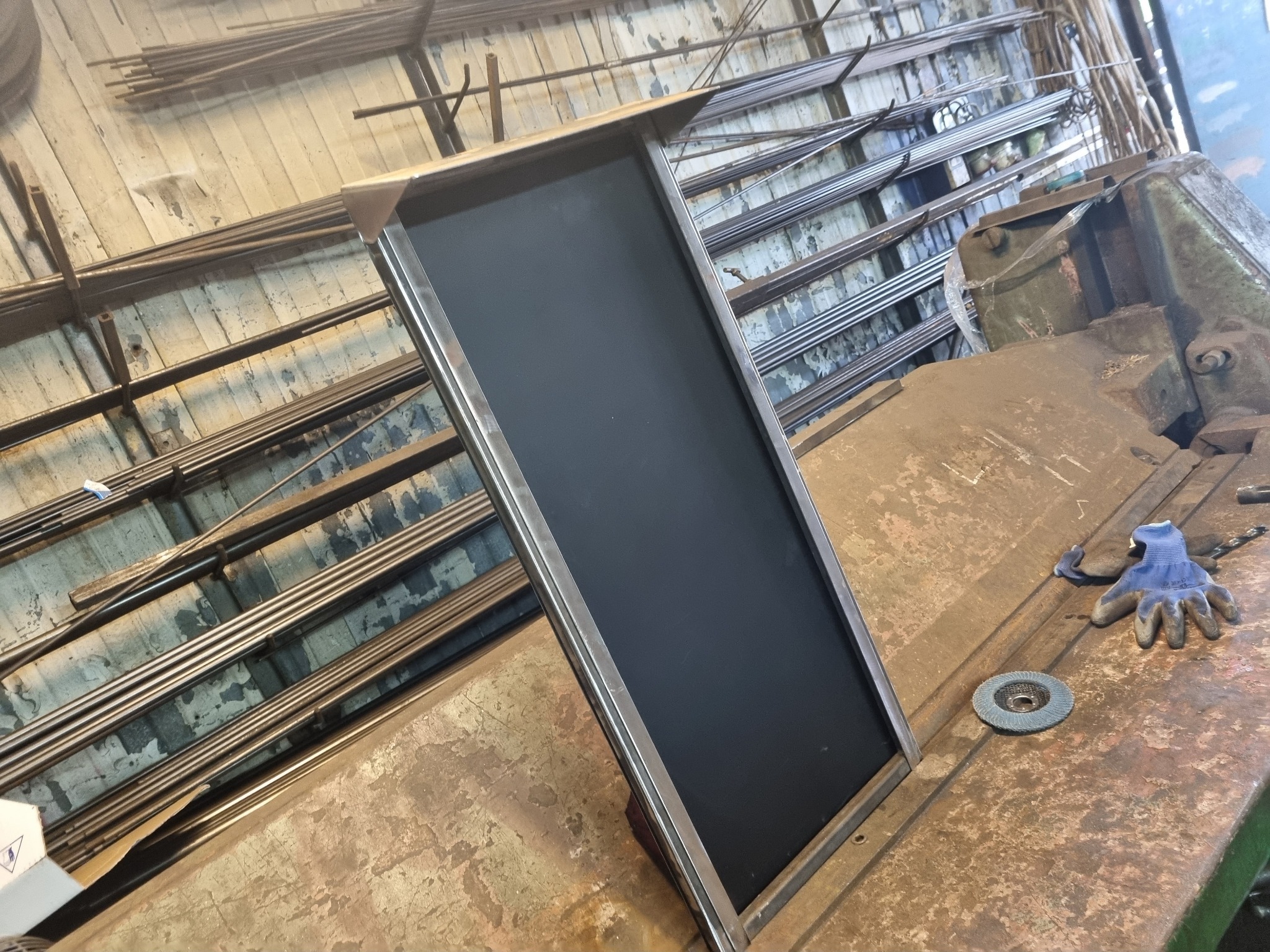 Metalowa rama z czarnym wypełnieniem na warsztatowym stole, obok tarcza szlifierska i rękawice robocze, w tle składowane metalowe pręty.