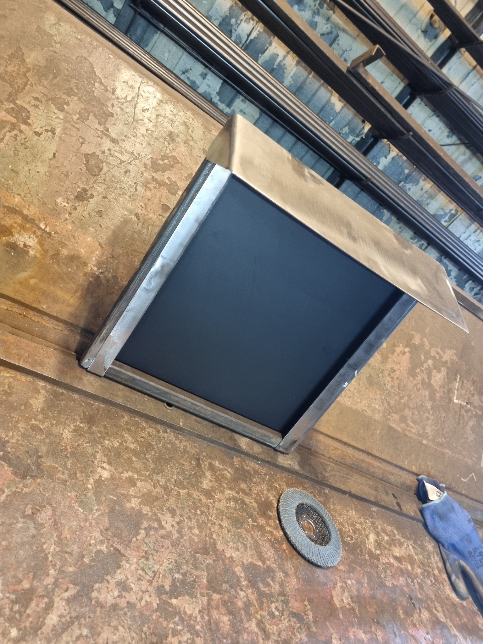 Surowy metalowy element z czarną wstawką, prawdopodobnie fragment konstrukcji, leżący na zardzewiałym metalowym stole warsztatowym w pobliżu tarczy szlifierskiej i rękawicy roboczej.