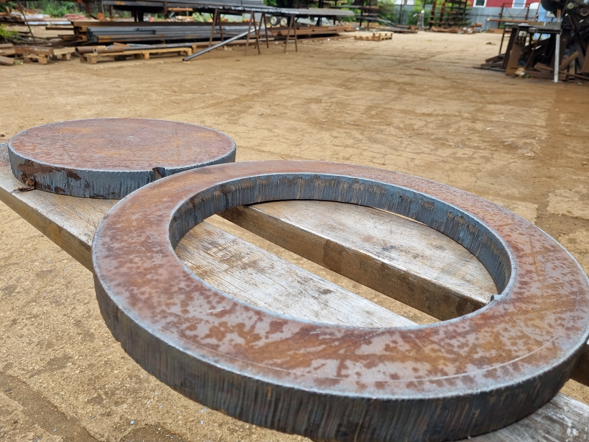 Dwa surowe, metalowe elementy: okrągły pierścień i pełne koło, leżące na drewnianej palecie w warsztacie, widoczne ślady obróbki i rdzy.