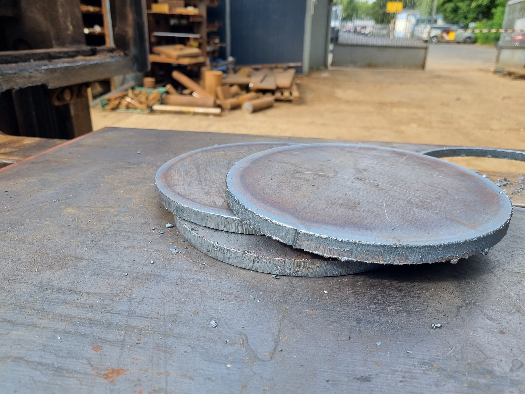 Trzy metalowe dyski o surowych krawędziach, ułożone jeden na drugim na metalowym stole warsztatowym, z widocznymi śladami obróbki i drobnymi opiłkami.