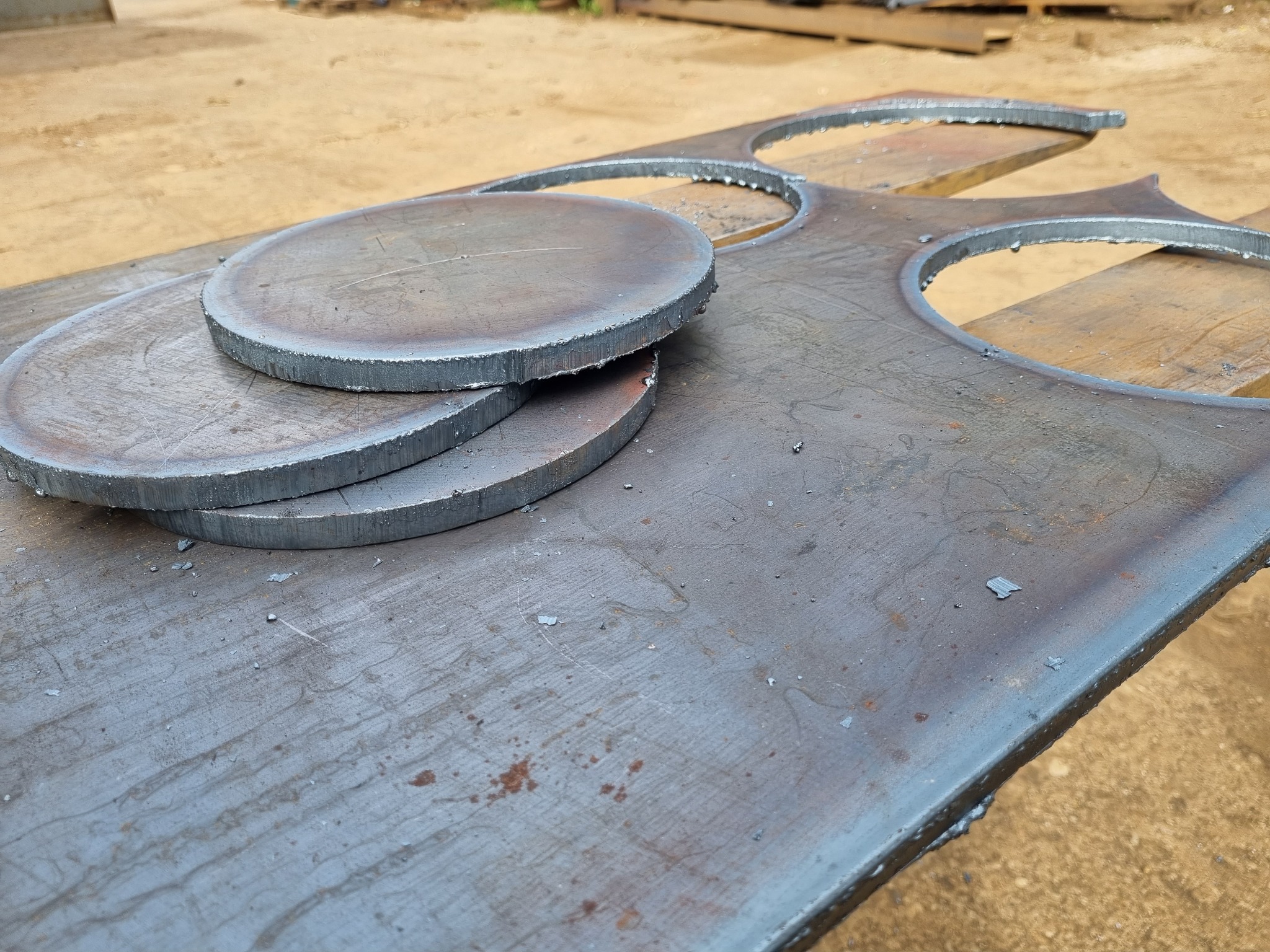 Stos trzech okrągłych, wyciętych metalowych elementów leżących na metalowej płycie z wyciętymi otworami, na drewnianym stole, widoczne ślady obróbki.