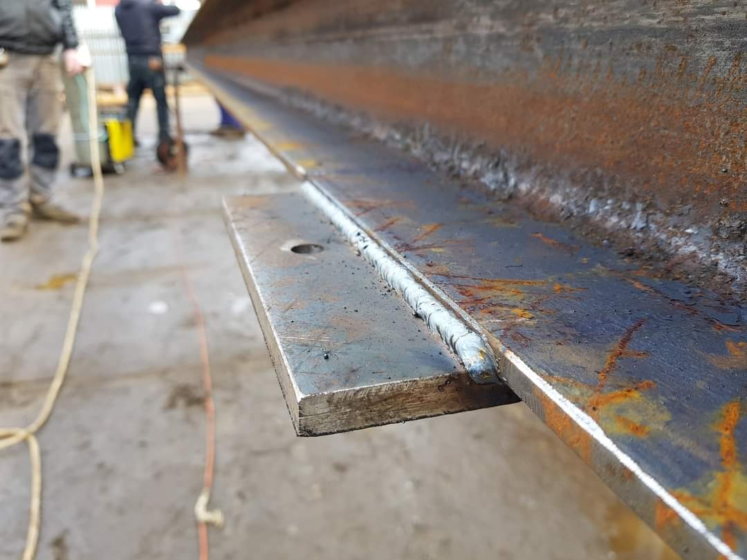 Spoina łącząca płaskownik z rdzewiejącą, stalową belką dwuteową, widoczne osoby i sprzęt spawalniczy w tle.