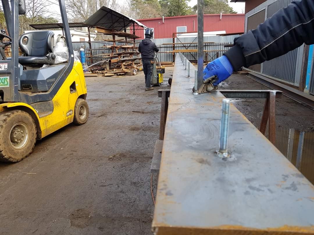 Pracownik w Józefowie czyści spoinę na długim, metalowym profilu z wystającymi śrubami, w tle wózek widłowy i plac budowy.