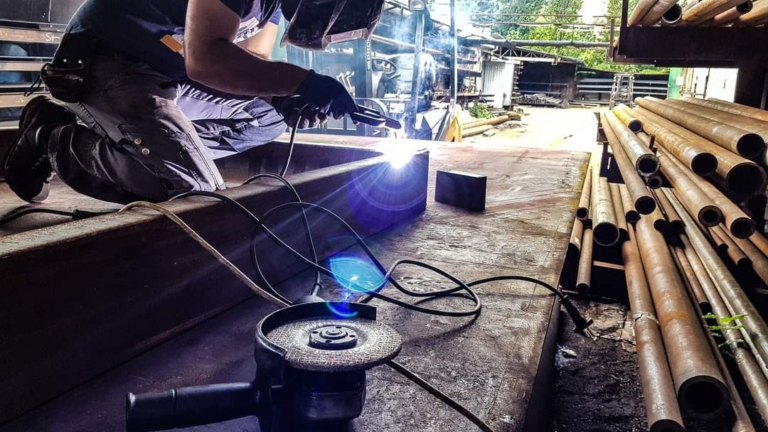 Spawanie metalowego profilu przez spawacza w ochronnej masce, widoczne iskry i narzędzia, w tle rury i konstrukcja hali.