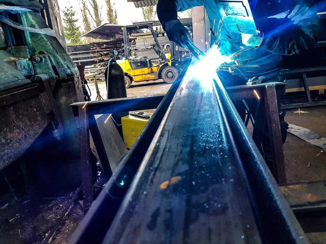 Praca w warsztacie: spawanie elementu metalowego o profilu ceownika, widoczne iskry, spawarka, butla z gazem i wózek widłowy w tle.