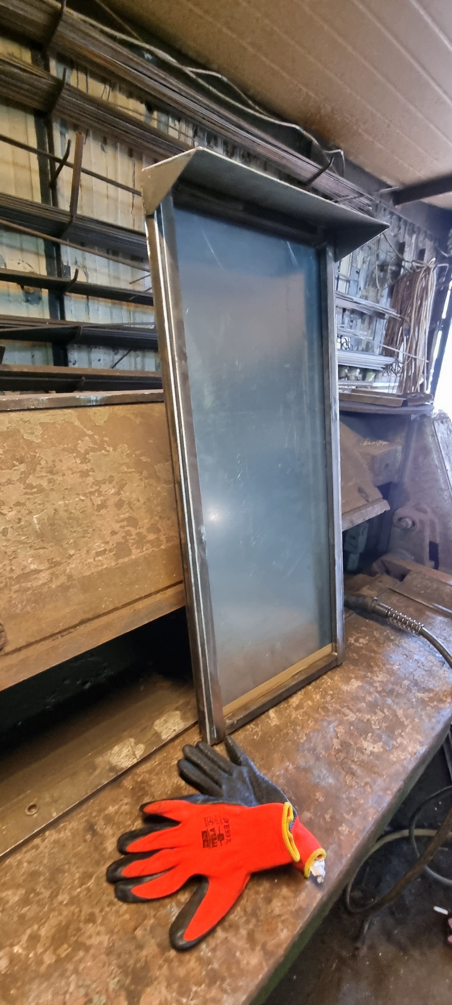 Metalowa rama z blachą wewnątrz, leżąca na warsztatowym stole, obok para czerwonych rękawic roboczych, w tle półki z metalowymi profilami.