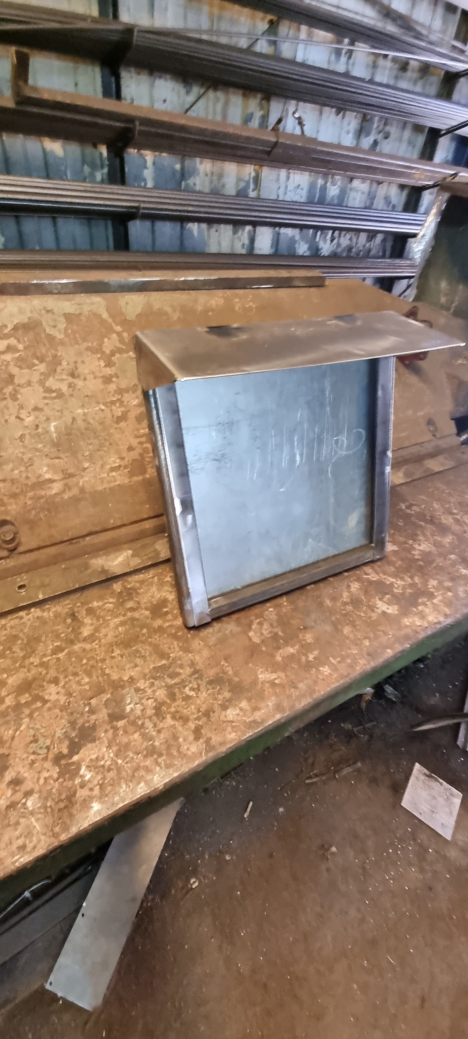 Metalowa konstrukcja z blachy, prawdopodobnie obudowa lub skrzynka, z widocznymi śladami po pisaniu markerem, leżąca na zardzewiałym metalowym stole w warsztacie.