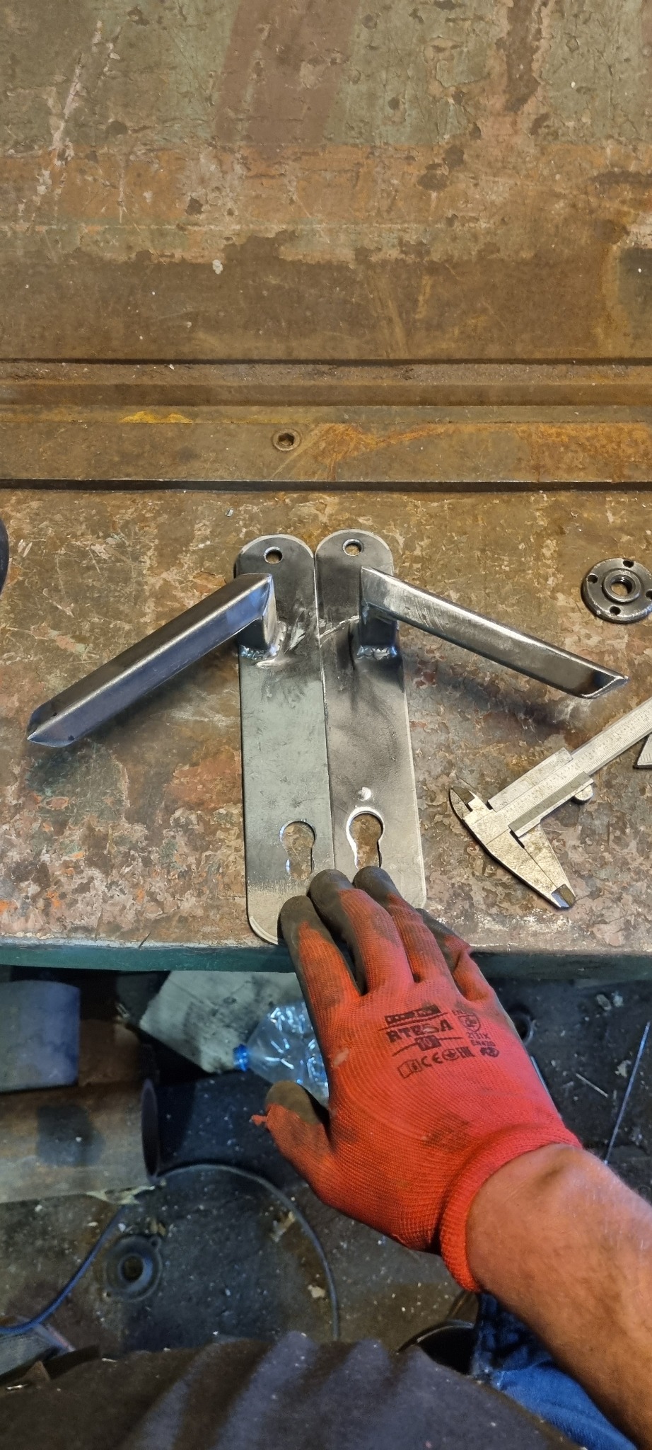 Dwie surowe, metalowe klamki do drzwi na warsztatowym stole, obok suwmiarka i dłoń w roboczej rękawicy.