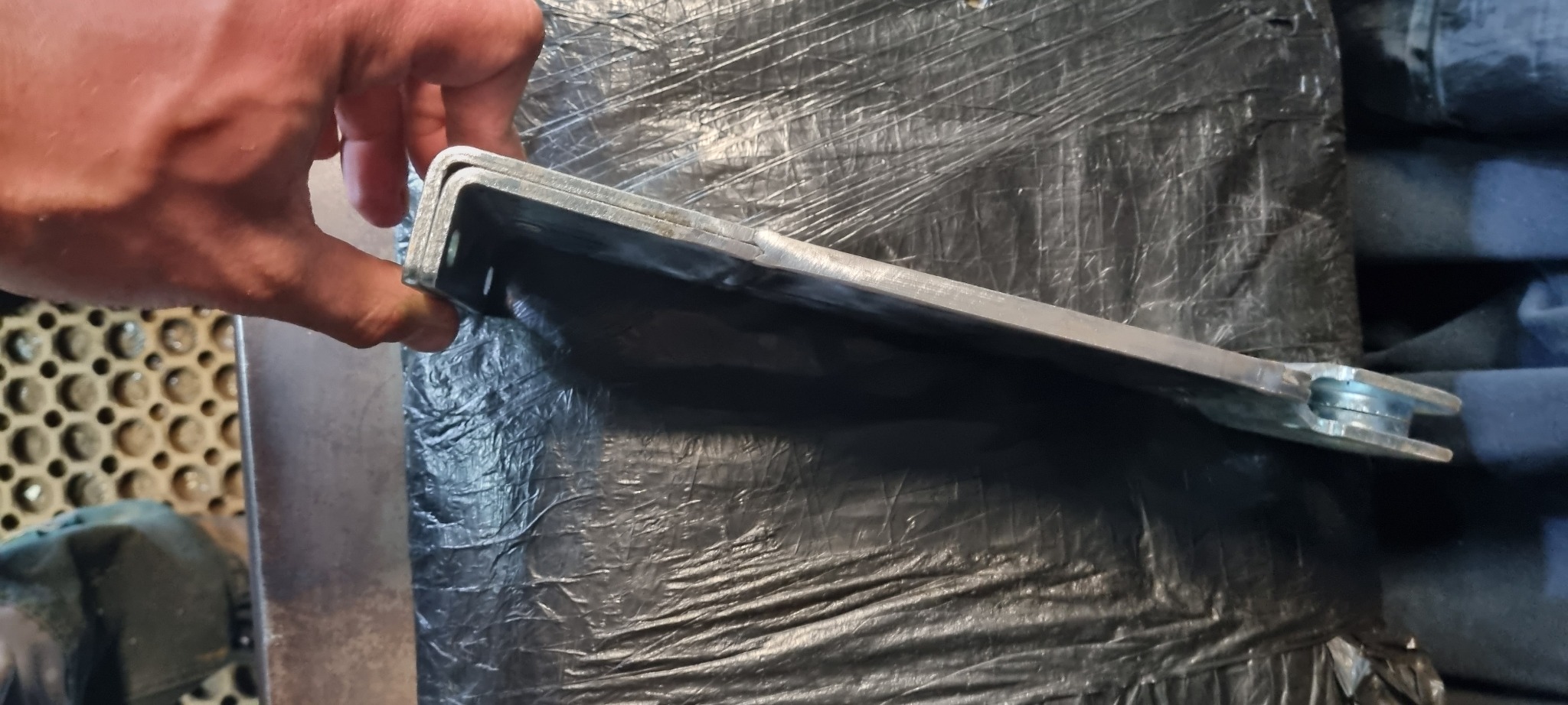 Profil stalowy kątowy z otworami, trzymany w dłoni, na tle ciemnej folii i fragmentu podłogi warsztatowej.