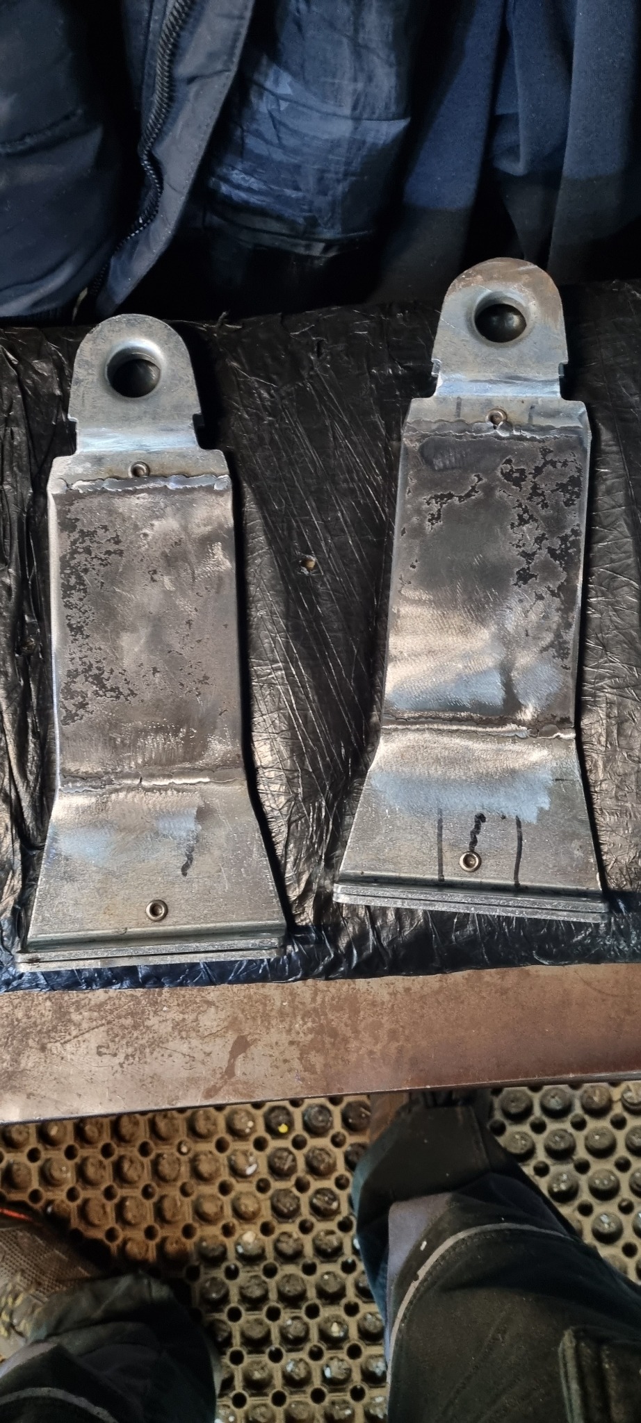 Dwa metalowe elementy z otworem i śladami spawania leżące na czarnej folii, widoczne ślady obróbki.