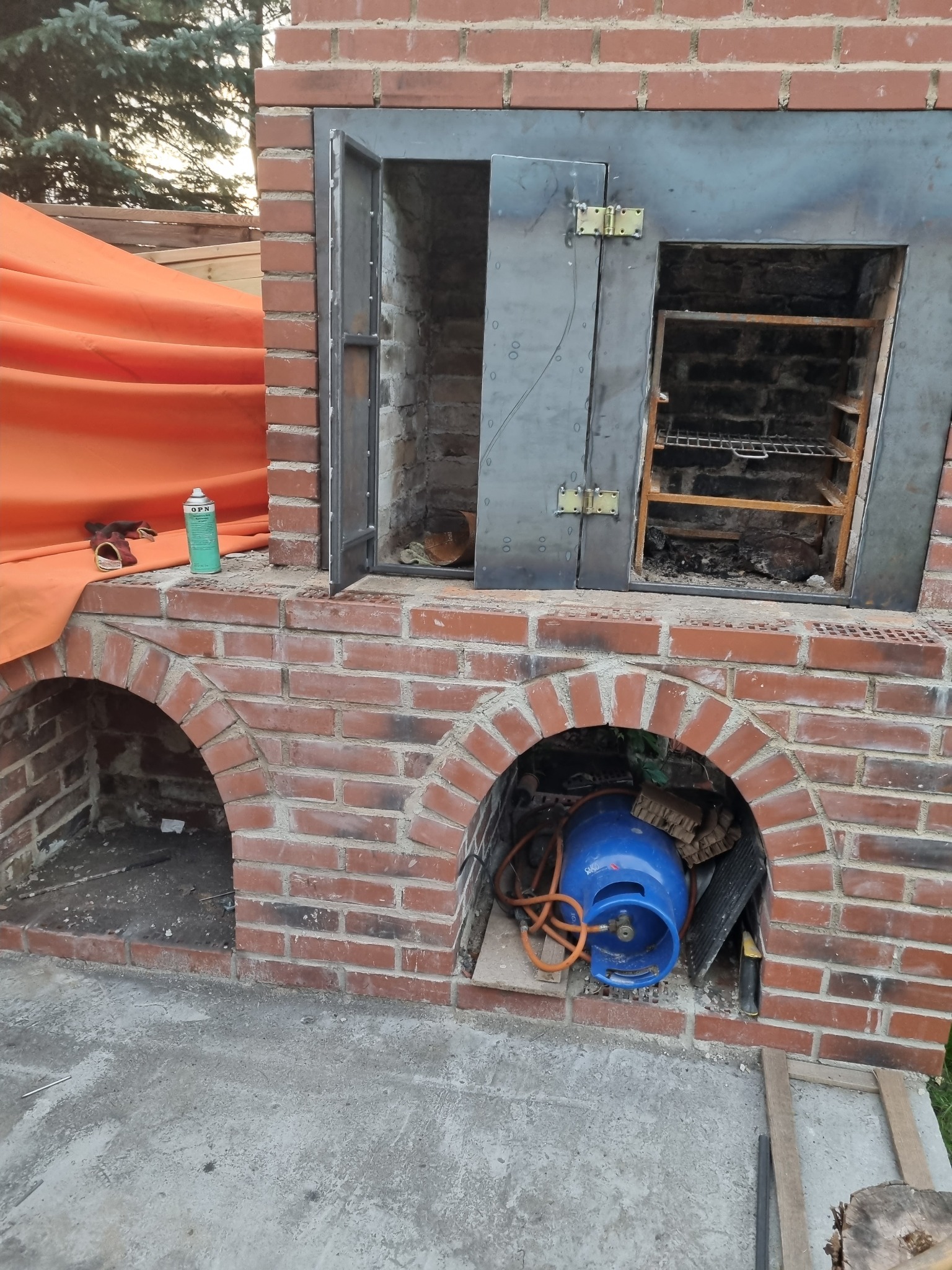 Murowany grill ogrodowy z metalowymi drzwiczkami i rusztem, częściowo zakryty pomarańczową tkaniną, z butlą gazową i narzędziami w pobliżu.