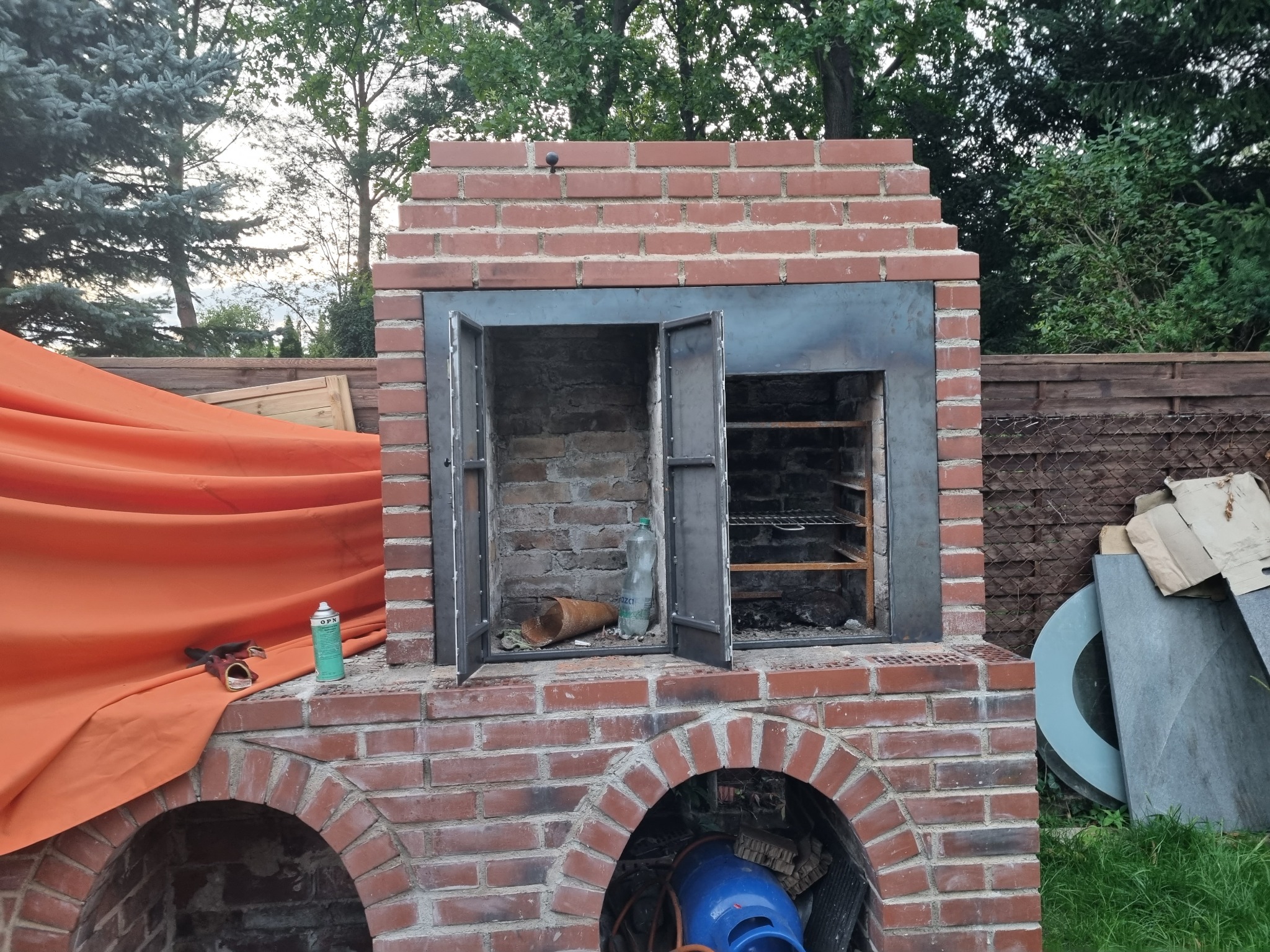 Murowany grill z metalowymi drzwiczkami w trakcie budowy, z otwartymi drzwiami, widoczne narzędzia i materiały budowlane, pomarańczowa plandeka w tle.
