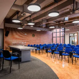 Sala konferencyjna z rzędami niebieskich krzeseł, oświetleniem sufitowym z okrągłymi lampami i ceglaną ścianą w tle.