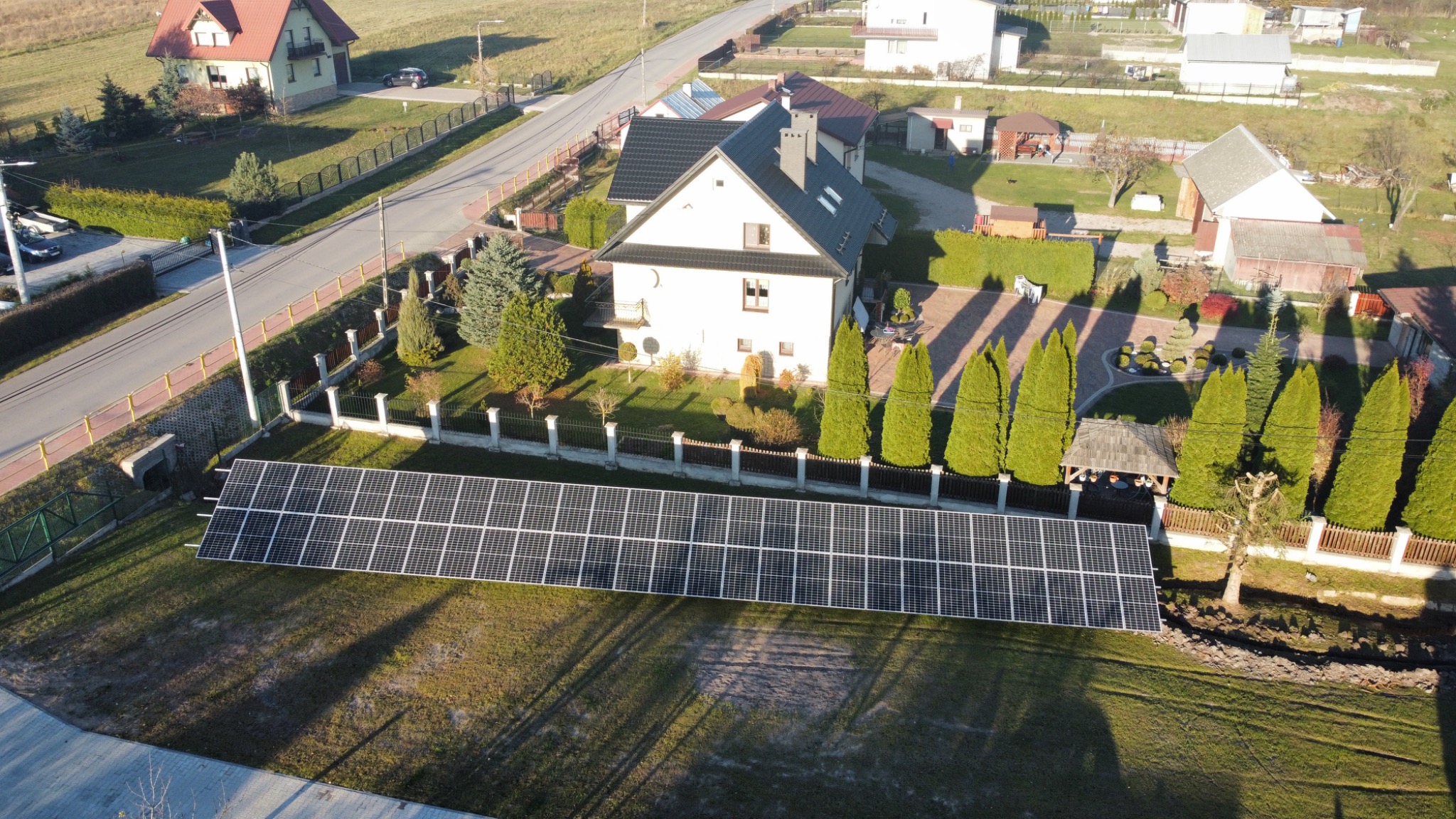Widok z lotu ptaka na instalację paneli słonecznych na trawniku przed domem jednorodzinnym z ciemnym dachem i zadbanym ogrodem, w tle inne budynki mieszkalne i droga.