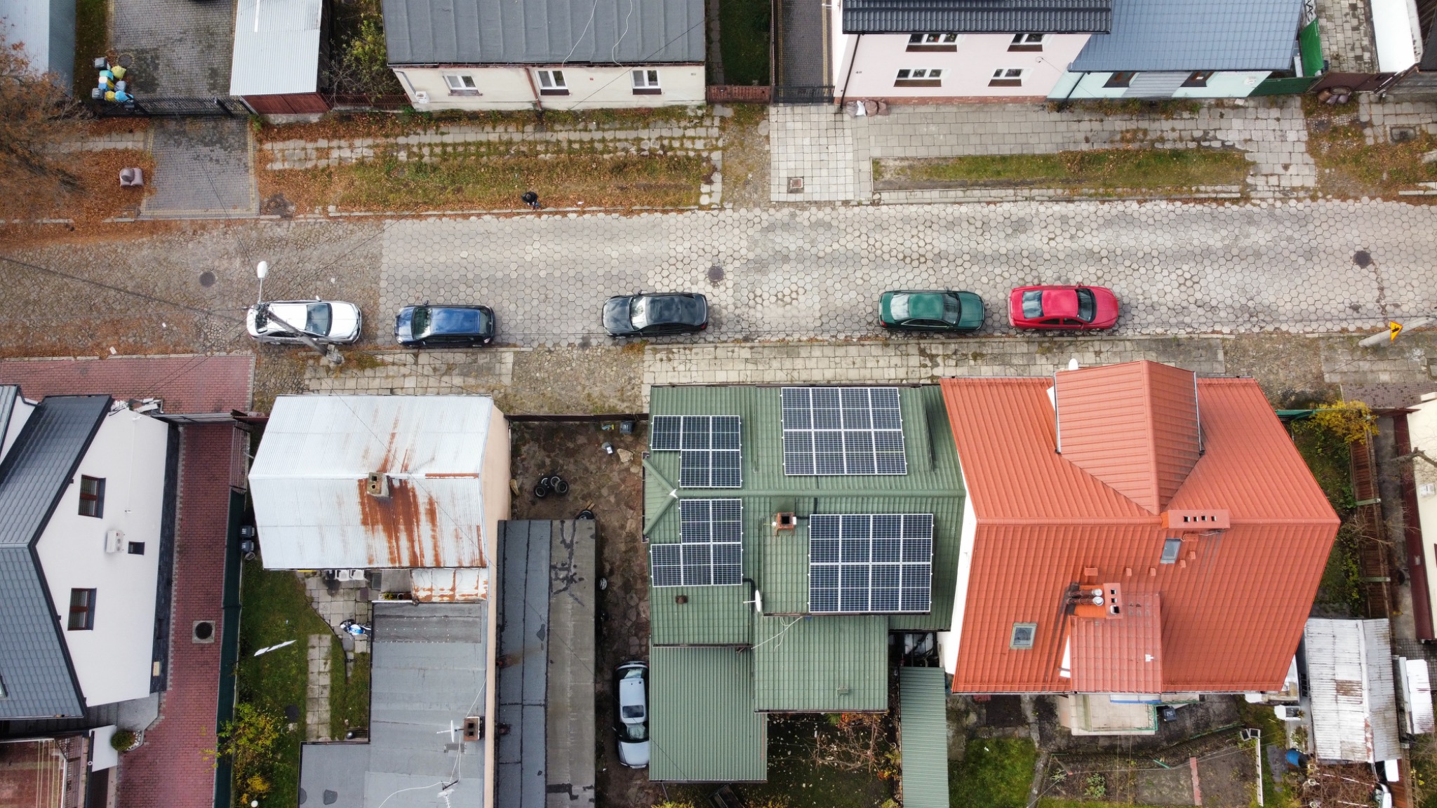 Widok z drona na dom z panelami fotowoltaicznymi na zielonym dachu, zaparkowane samochody wzdłuż ulicy z kostki brukowej, sąsiednie budynki z czerwonymi dachami.