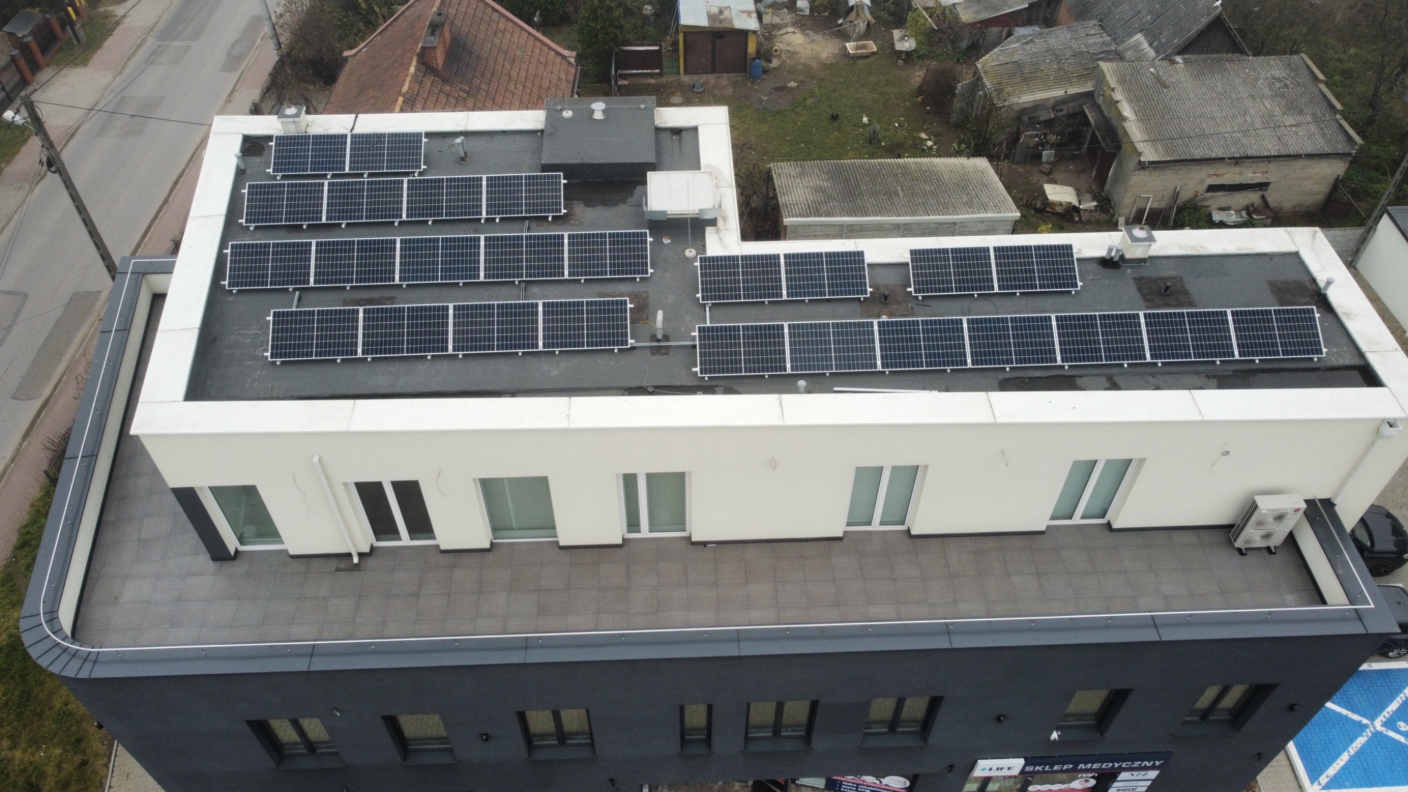 Widok z góry na budynek z zainstalowanymi panelami słonecznymi na płaskim dachu, widoczny fragment ulicy i zabudowań w tle.