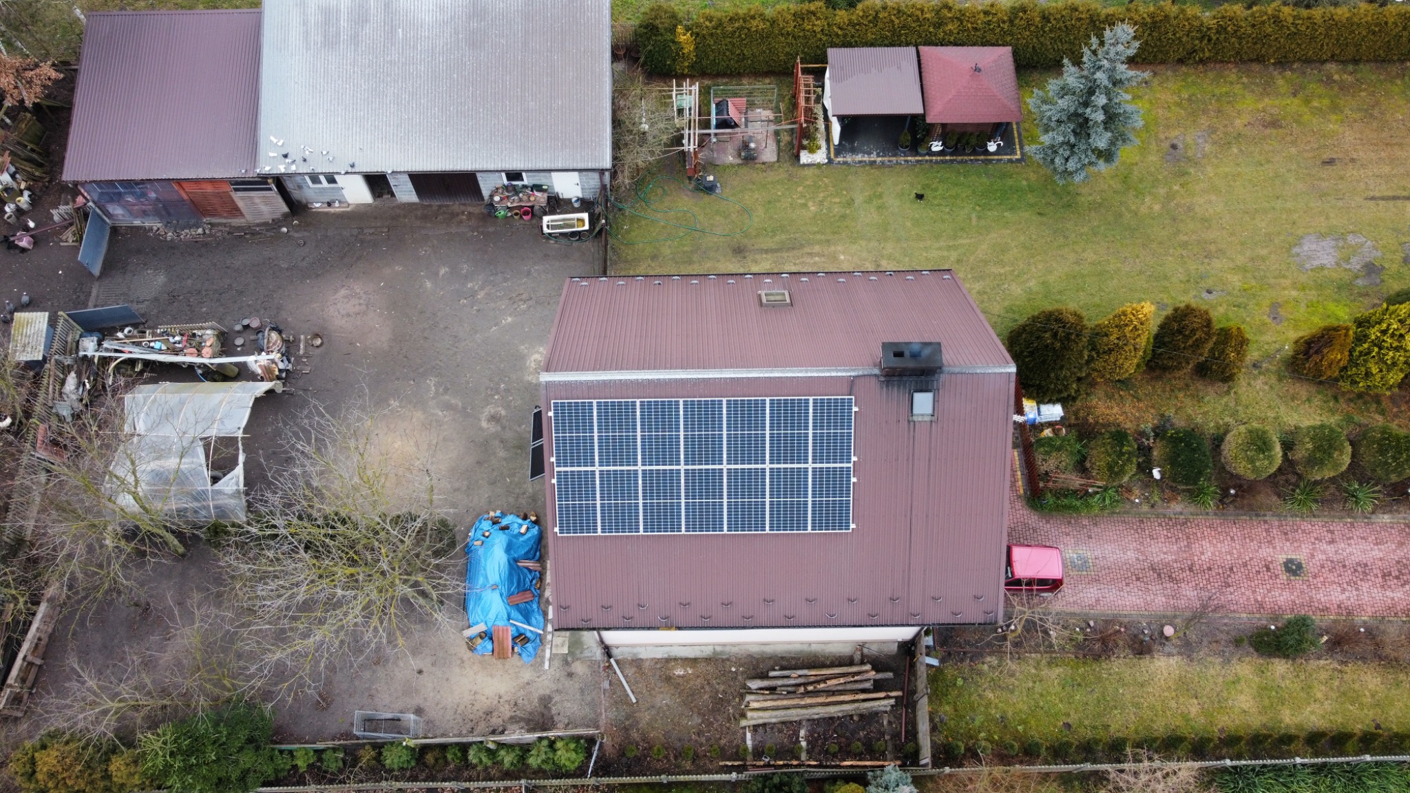 Widok z góry na dom z brązowym dachem, na którym zamontowano panele słoneczne, obok budynki gospodarcze i ogród.