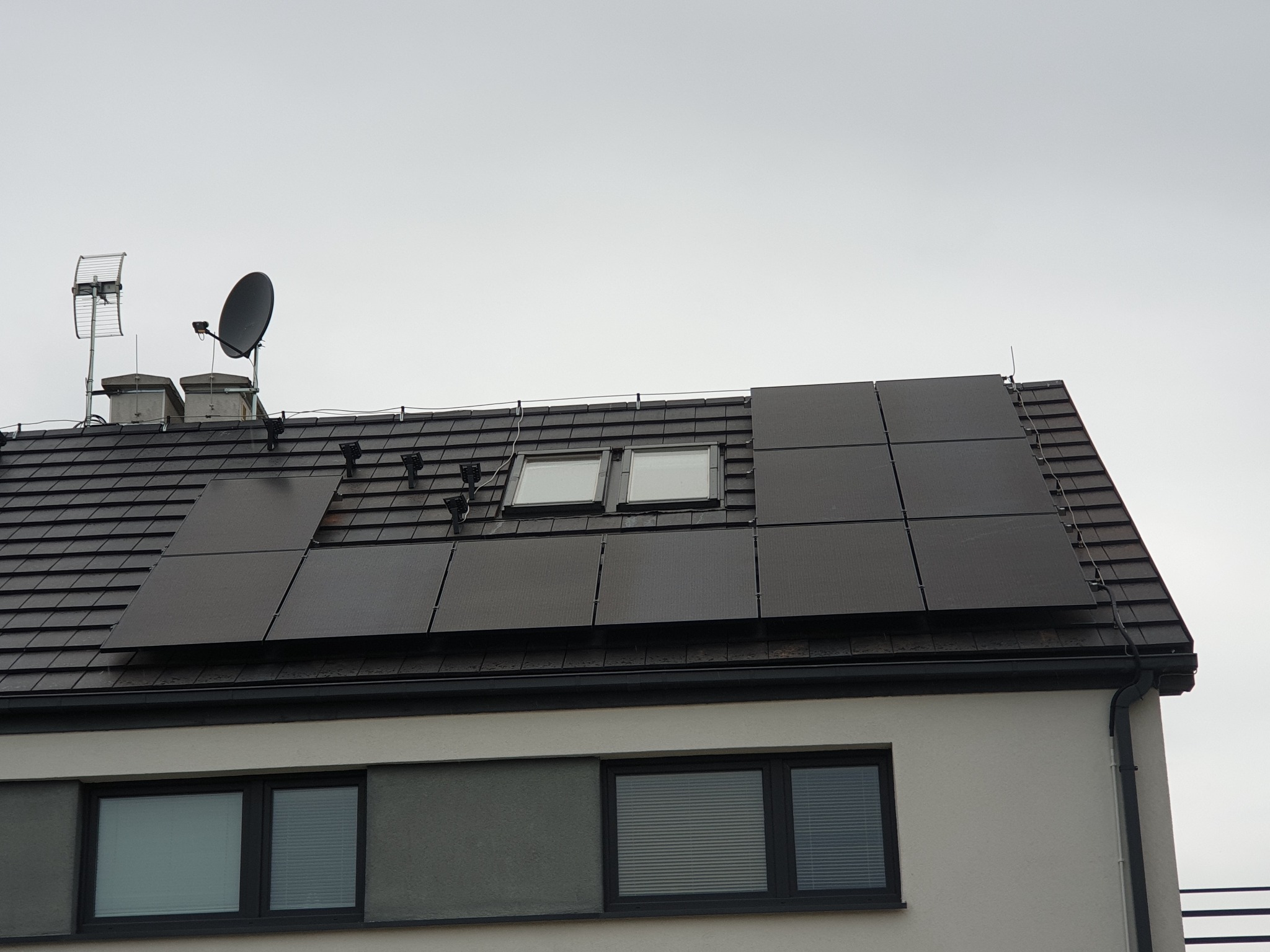 Częściowy widok domu z zainstalowanymi panelami fotowoltaicznymi na dachu z dachówki ceramicznej, anteną satelitarną i telewizyjną oraz oknami dachowymi.
