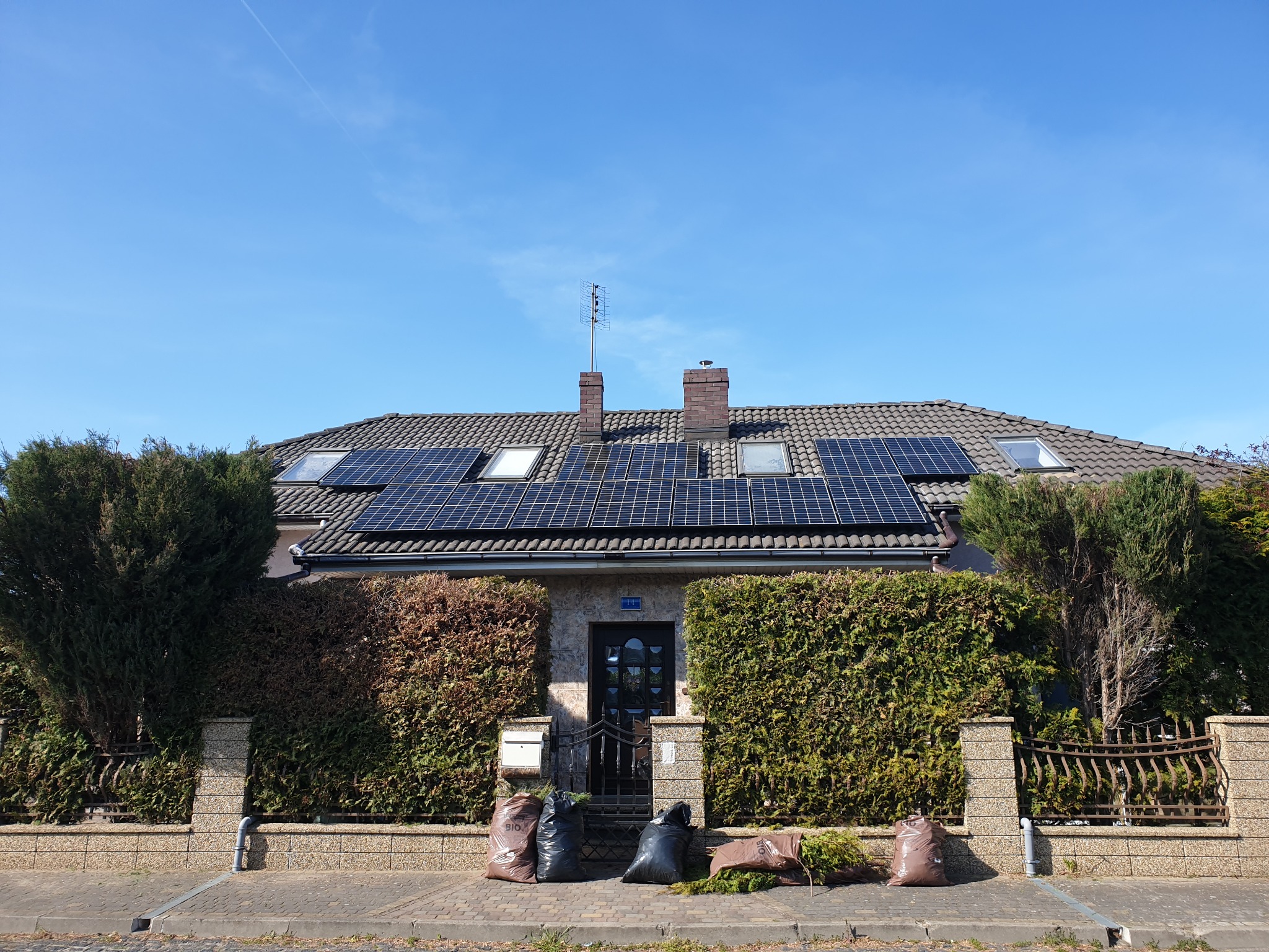 Dom jednorodzinny z panelami słonecznymi na dachu, widoczne również okna dachowe, antena telewizyjna, żywopłot i worki z odpadami ogrodowymi przed wejściem.