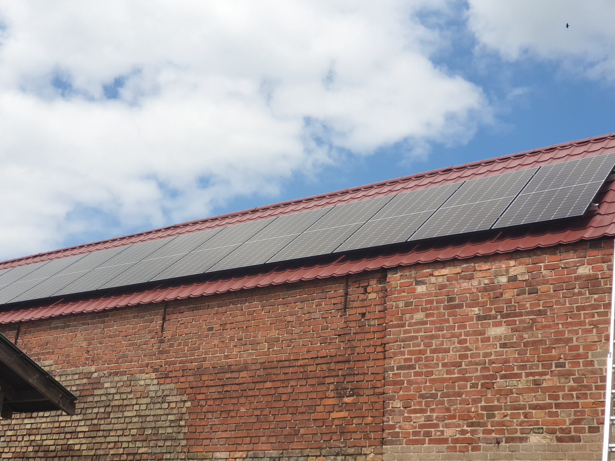 Instalacja paneli słonecznych na czerwonym, ceglanym budynku we Wrocławiu, widok z dołu na dach z panelami i fragment drabiny po prawej stronie.