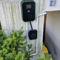 Czarna stacja ładowania pojazdów elektrycznych GC z zielonym obramowaniem, zamontowana na zewnętrznej ścianie budynku, obok rury i zielonego krzewu.