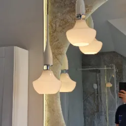 Dwa nowoczesne, białe wiszące oświetlenia w łazience z marmurowym wykończeniem, odbijające się w owalnym lustrze z podświetleniem LED, w tle widoczna kabina prysznicowa.