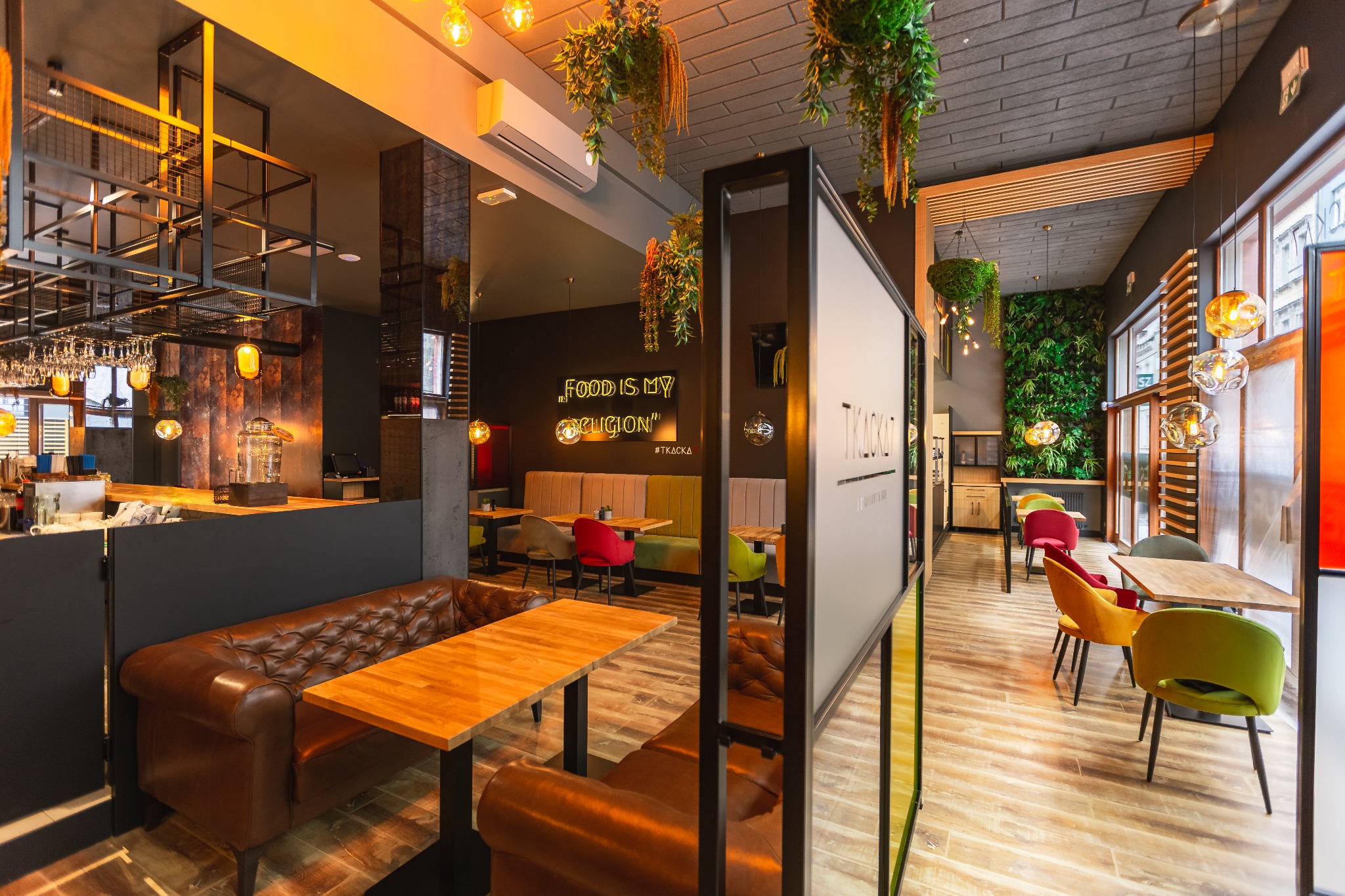 Wnętrze restauracji z zieloną ścianą, wiszącymi roślinami, skórzanymi kanapami i drewnianymi stołami, oświetlone ciepłym światłem żarówek.