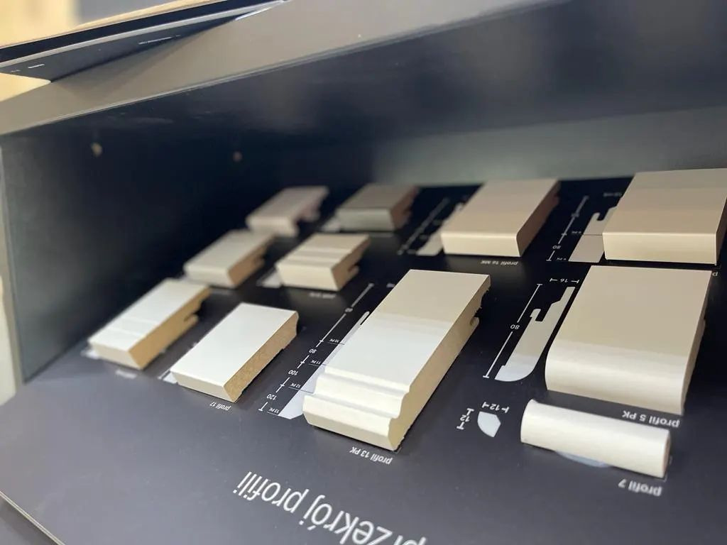 Ekspozycja próbek profili przypodłogowych o różnych kształtach i wymiarach, umieszczonych na czarnym tle z nadrukowanymi schematami przekrojów.