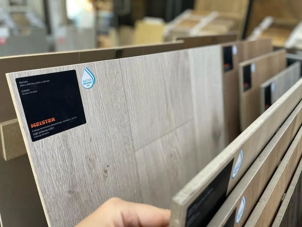 Próbki paneli podłogowych Meister, w odcieniach dębu polarnego, z etykietą informacyjną o wymiarach i odporności na wodę, prezentowane w ekspozytorze.