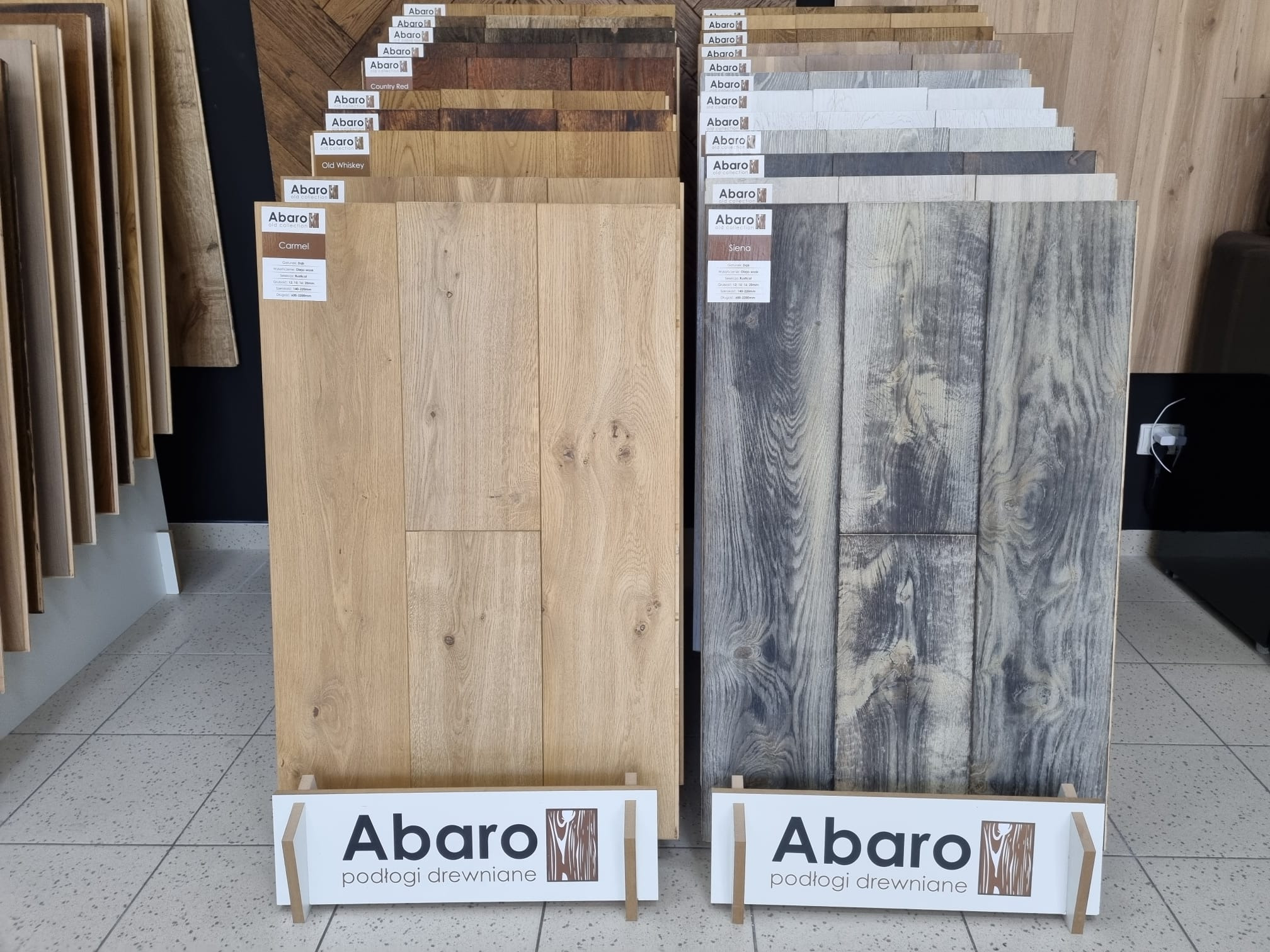 Ekspozycja próbek paneli podłogowych marki Abaro w różnych odcieniach: jasny dąb Carmel i szary Siena, z widocznym logo producenta.