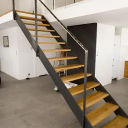 Nowoczesne schody z drewnianymi stopniami i metalową konstrukcją, widoczne z dołu, w tle minimalistyczne wnętrze z białymi ścianami i szklaną balustradą na antresoli.