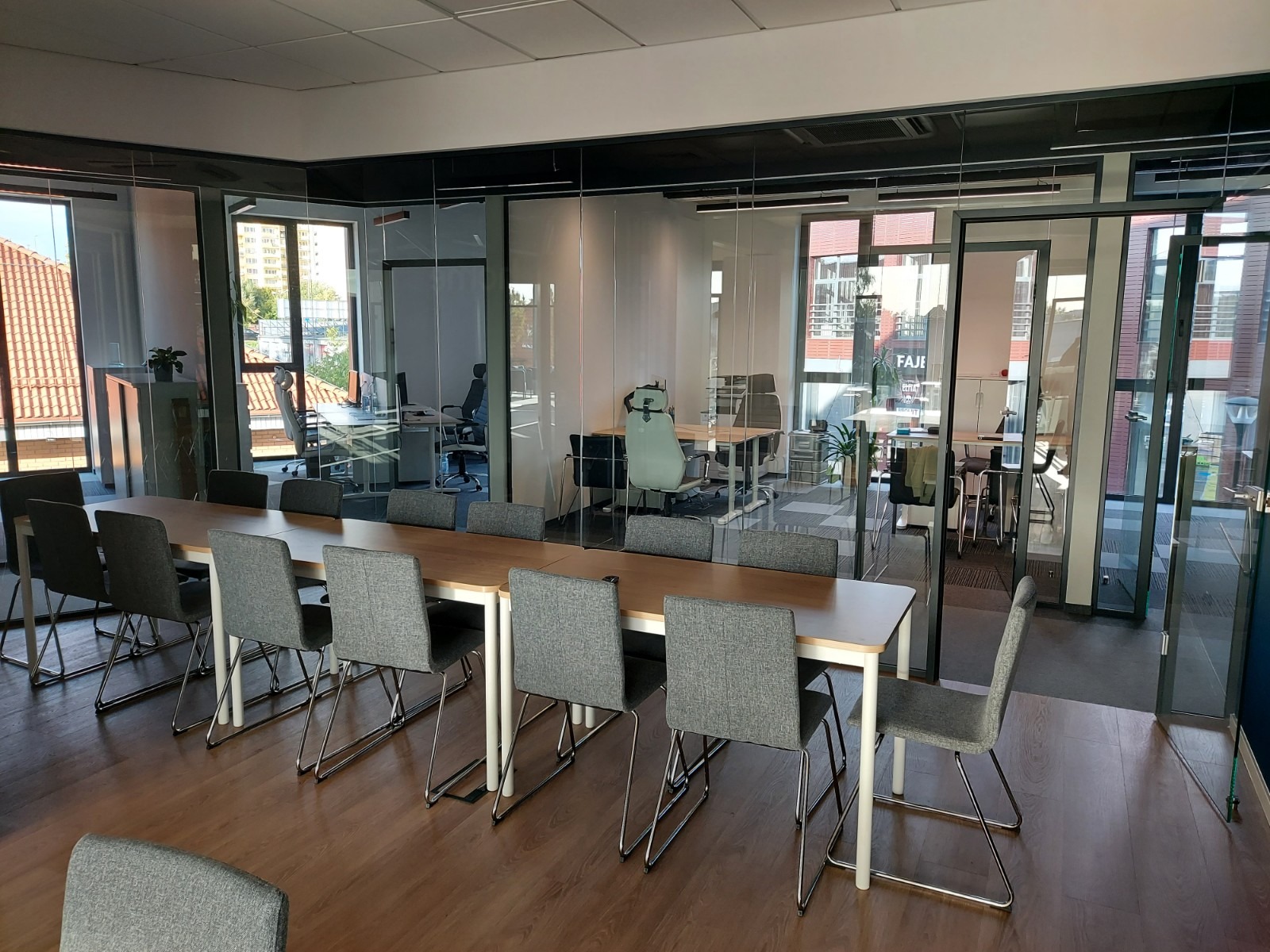 Nowoczesna sala konferencyjna z długim drewnianym stołem otoczonym szarymi krzesłami, oddzielona szklanymi ścianami od biur z widokiem na budynki z czerwonymi dachami.