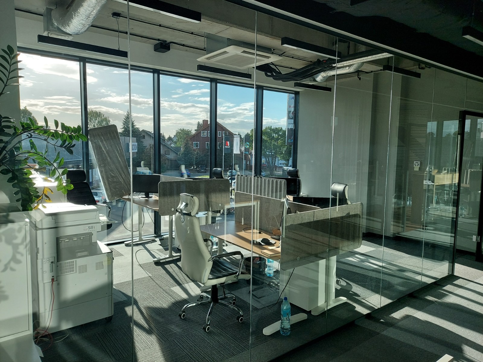 Nowoczesne biuro z przeszklonymi ścianami, widok na zewnątrz z domami, biurka z przegrodami, krzesła obrotowe, drukarka i butelka wody mineralnej.