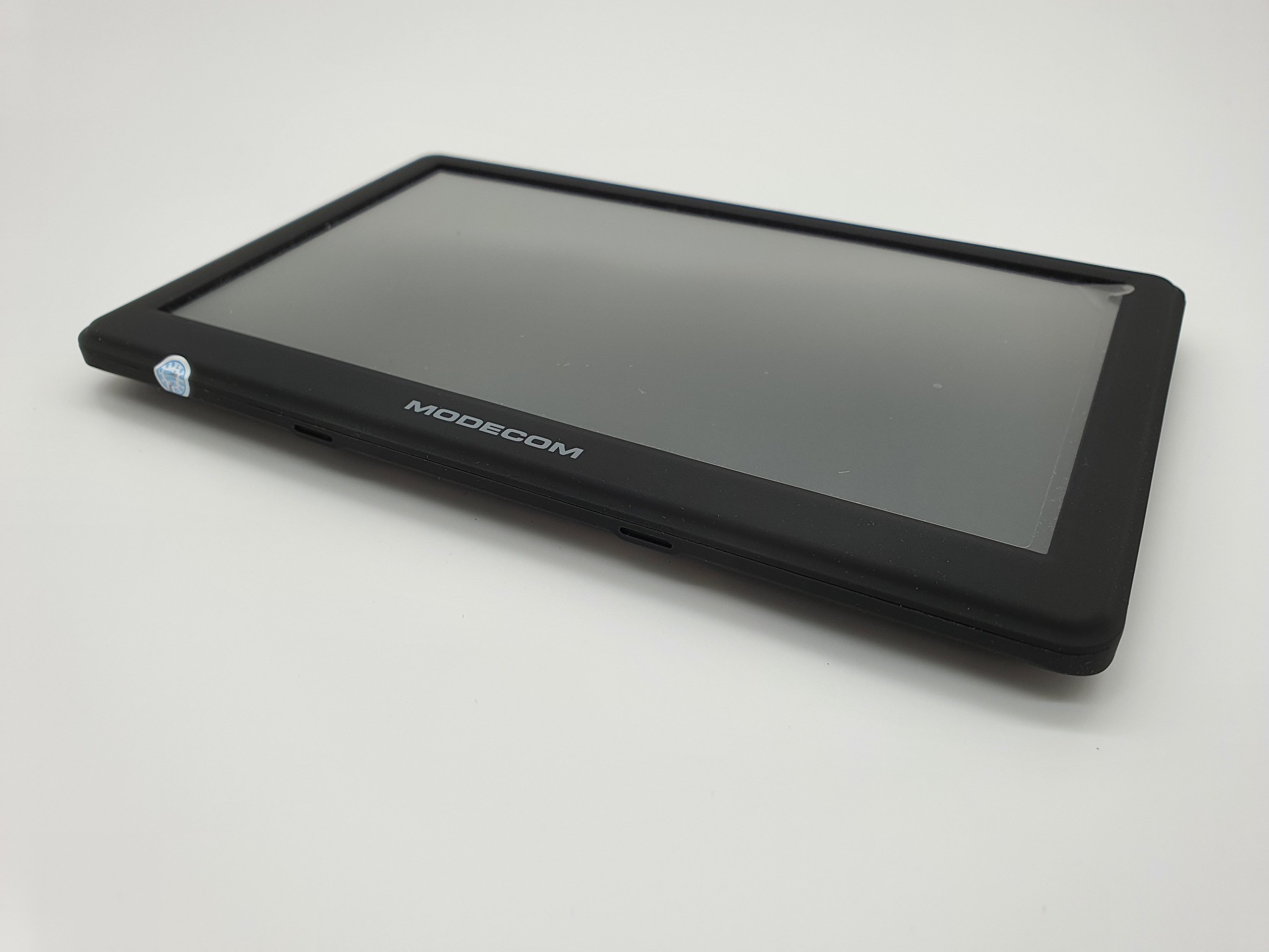 Czarny tablet marki Modecom leżący na białym tle, widoczna naklejka w lewym górnym rogu ekranu.