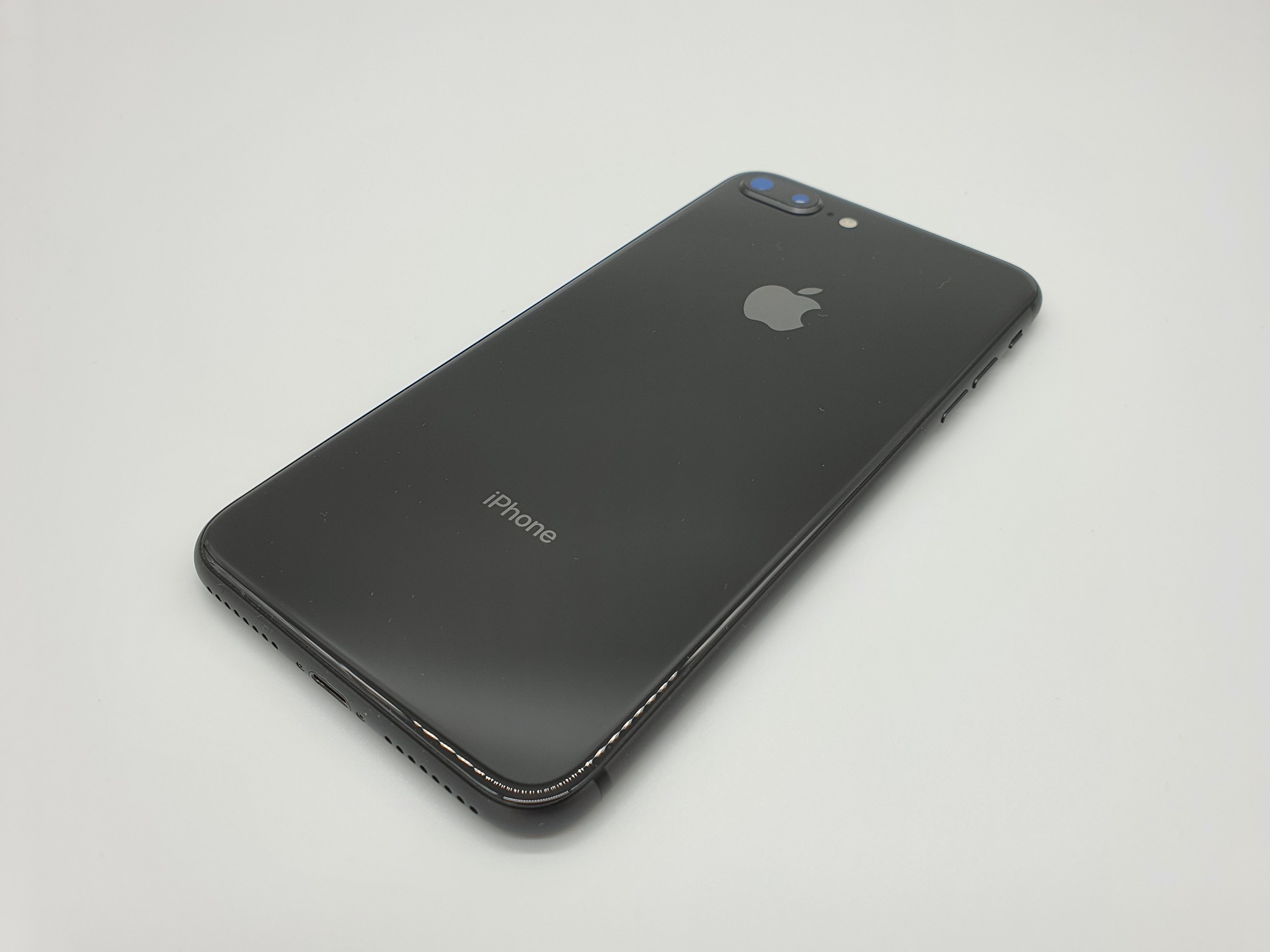 Czarny iPhone 7 Plus leżący na pleckach, widoczne logo Apple, podwójny obiektyw aparatu i napis iPhone, na jasnym tle.