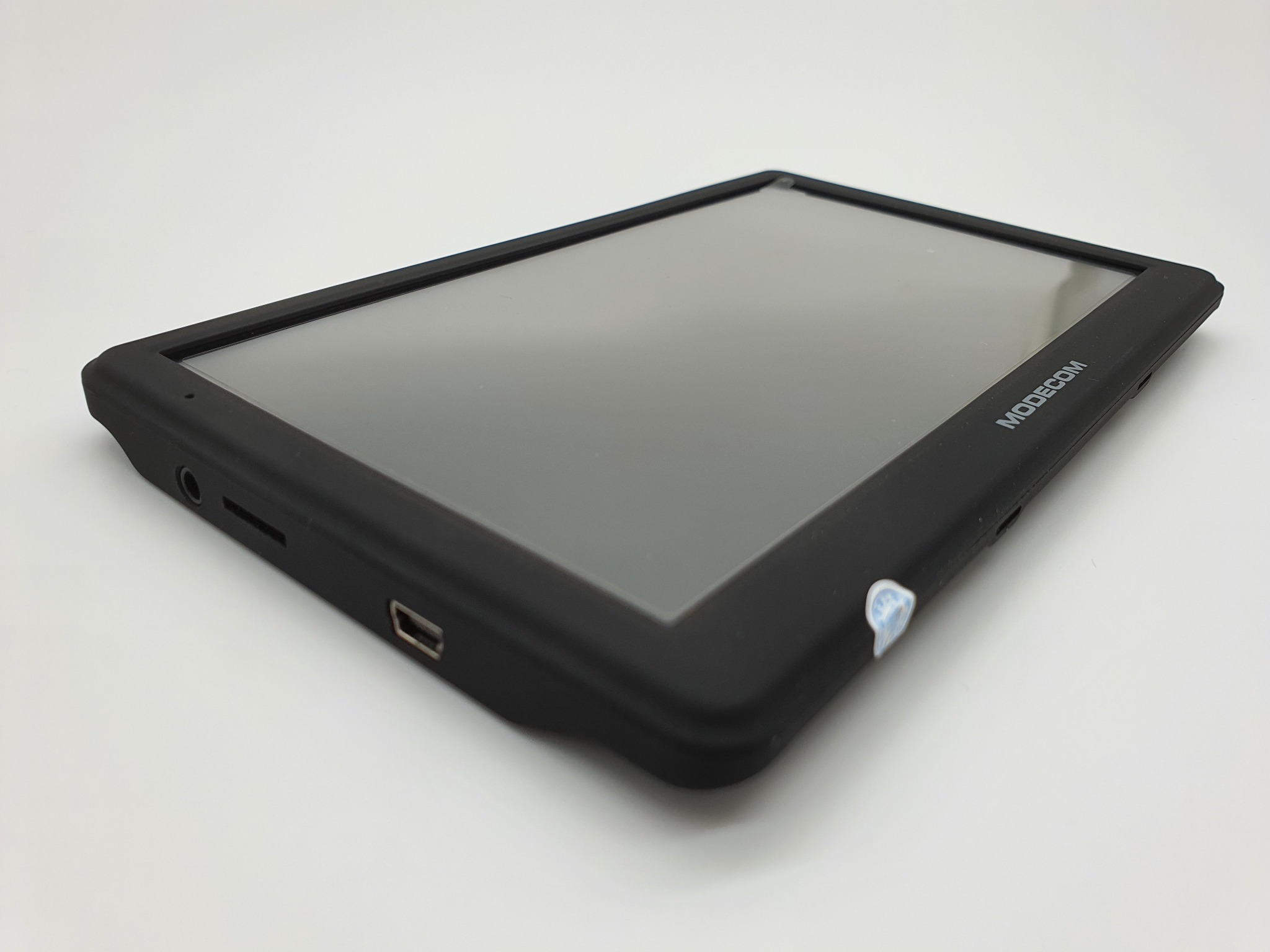 Tablet Modocom z czarną obudową, widoczne porty USB i słuchawkowe, na białym tle, z naklejką zabezpieczającą na obudowie.