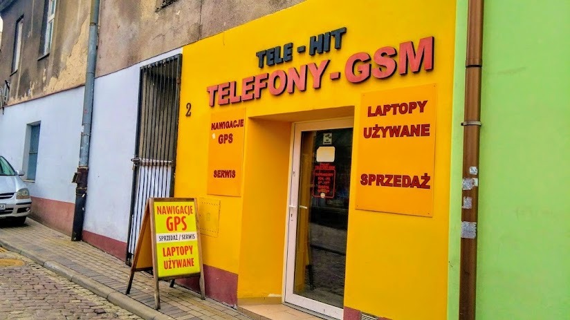 Fasada budynku z żółtą elewacją, szyldem 'TELE-HIT TELEFONY-GSM', tablicami informacyjnymi o nawigacjach GPS, laptopach używanych i sprzedaży, oraz stojakiem reklamowym z napisem 'Nawigacje GPS...
