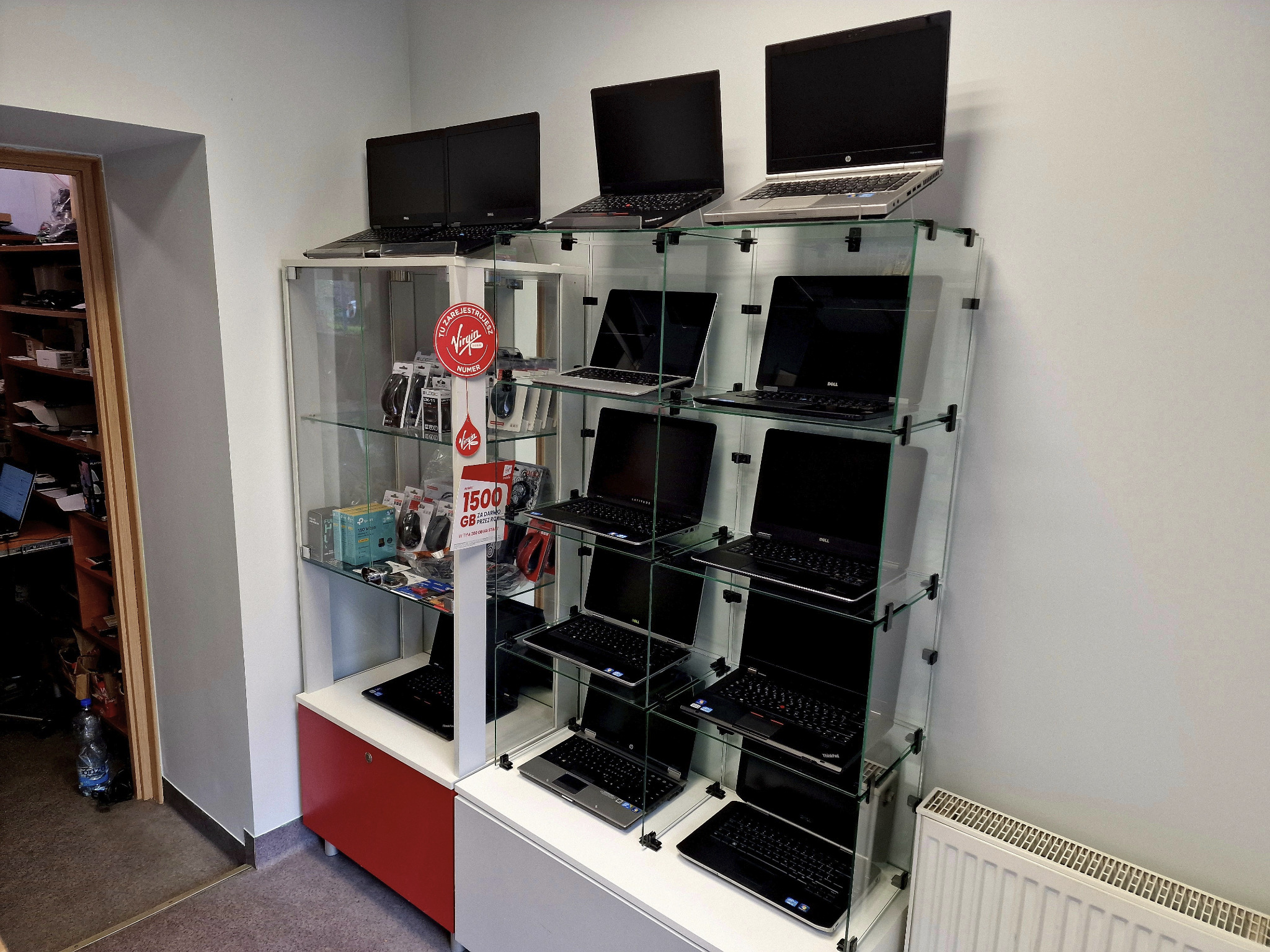 Ekspozycja używanych laptopów różnych marek (Dell, HP, ThinkPad) w szklanej witrynie w sklepie komputerowym. Widoczne również akcesoria komputerowe i reklama oferty specjalnej.